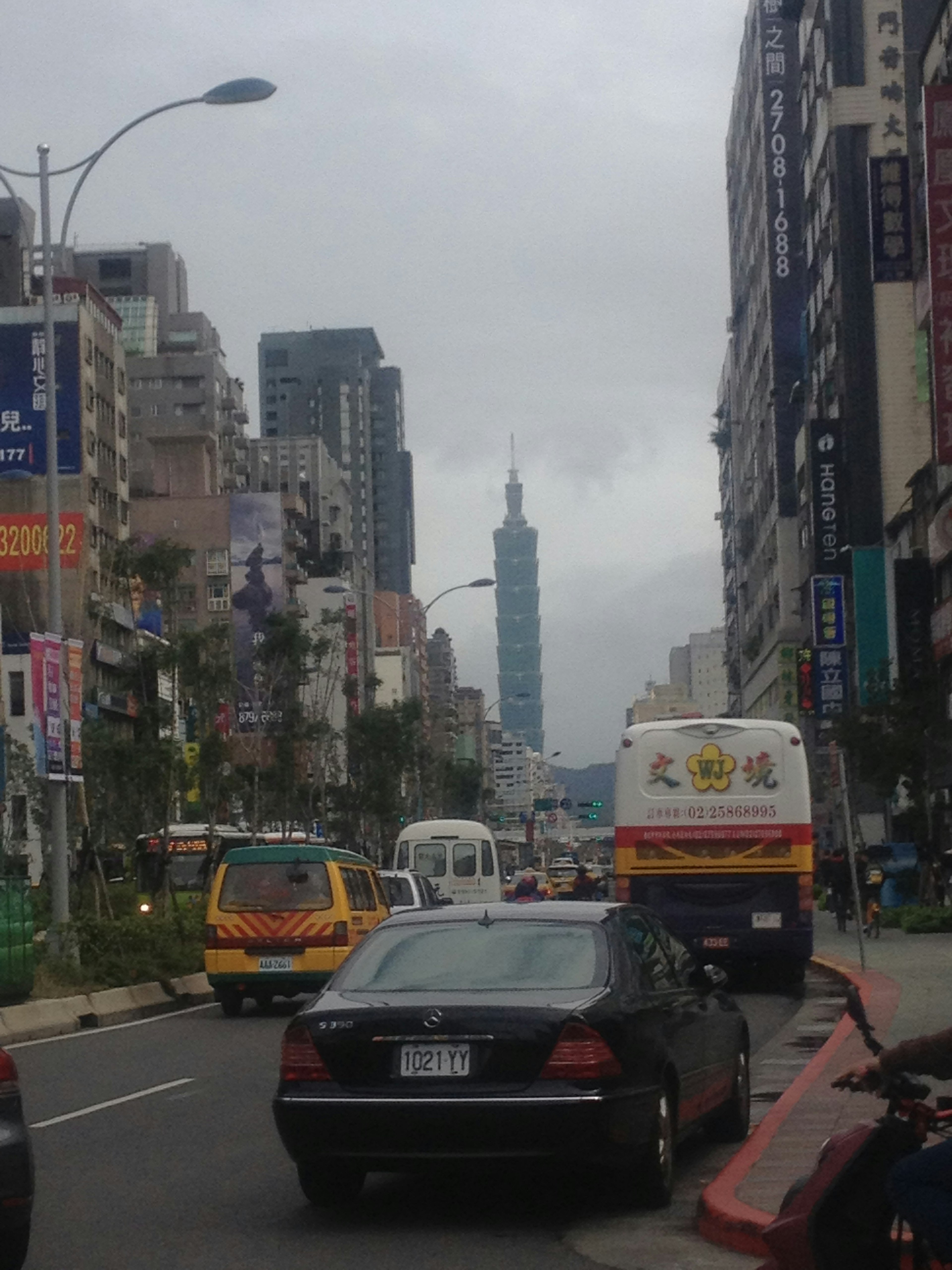 城市街道景觀，背景有台北101和汽車，天空多雲