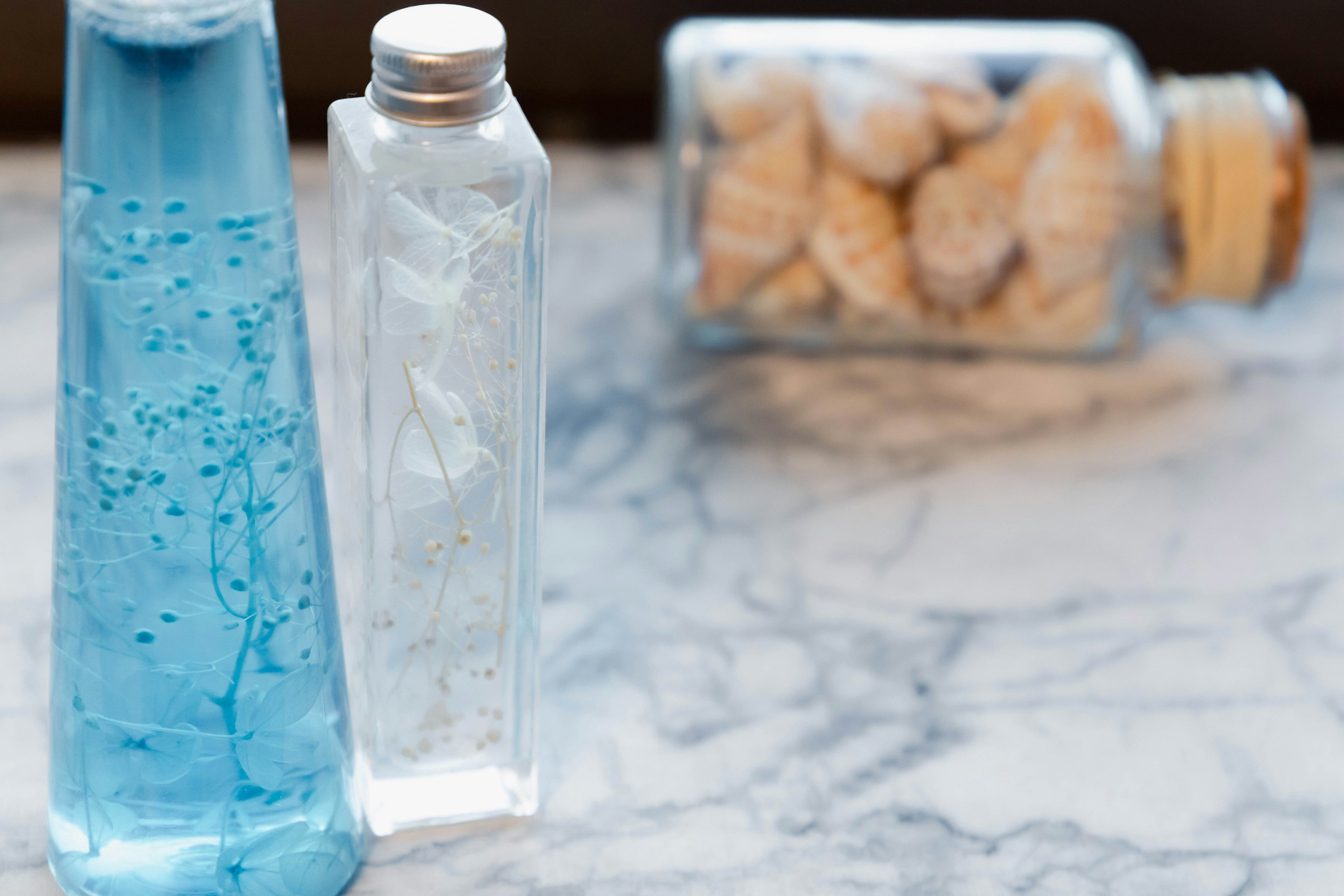 Dos botellas con líquido azul y líquido transparente con un frasco de conchas al fondo