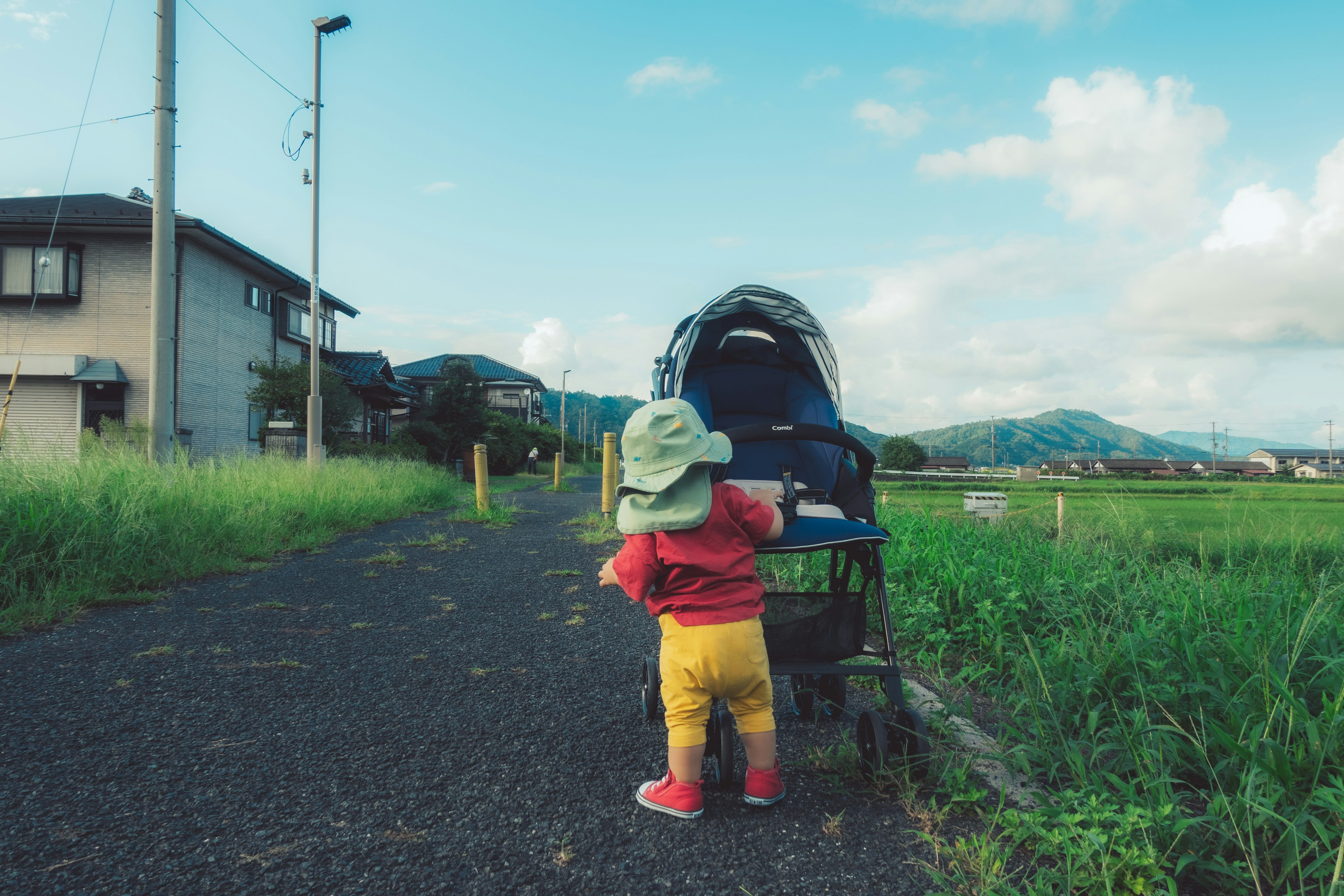 Un petit enfant se tenant devant une poussette sur un chemin rural avec de l'herbe verte et un ciel bleu