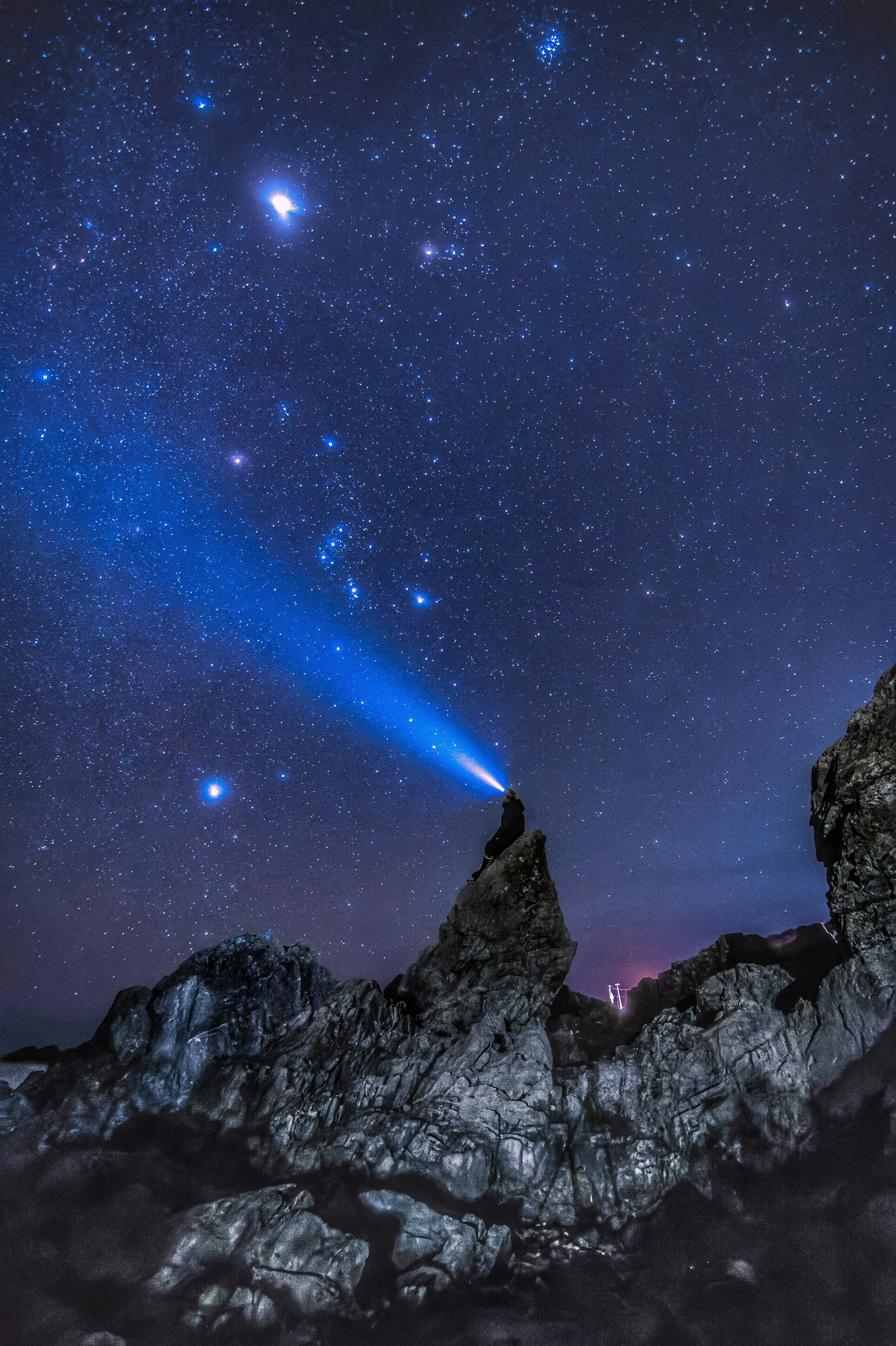 Una persona de pie en un pico rocoso bajo un cielo estrellado emitiendo un haz de luz