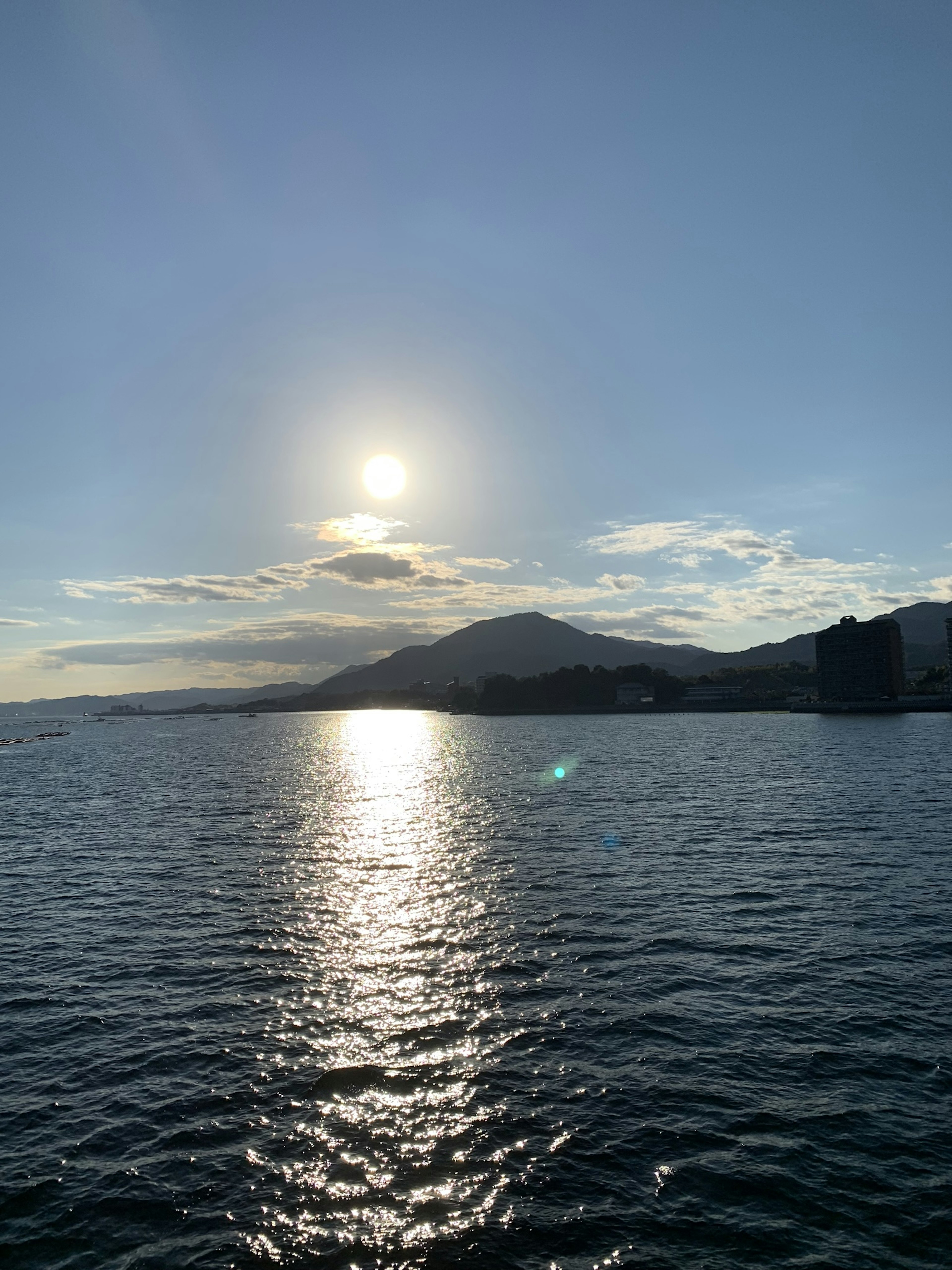 Malersiche Aussicht auf das Meer mit der Sonne, die in einem blauen Himmel scheint, Silhouette von Bergen und schimmerndem Wasser