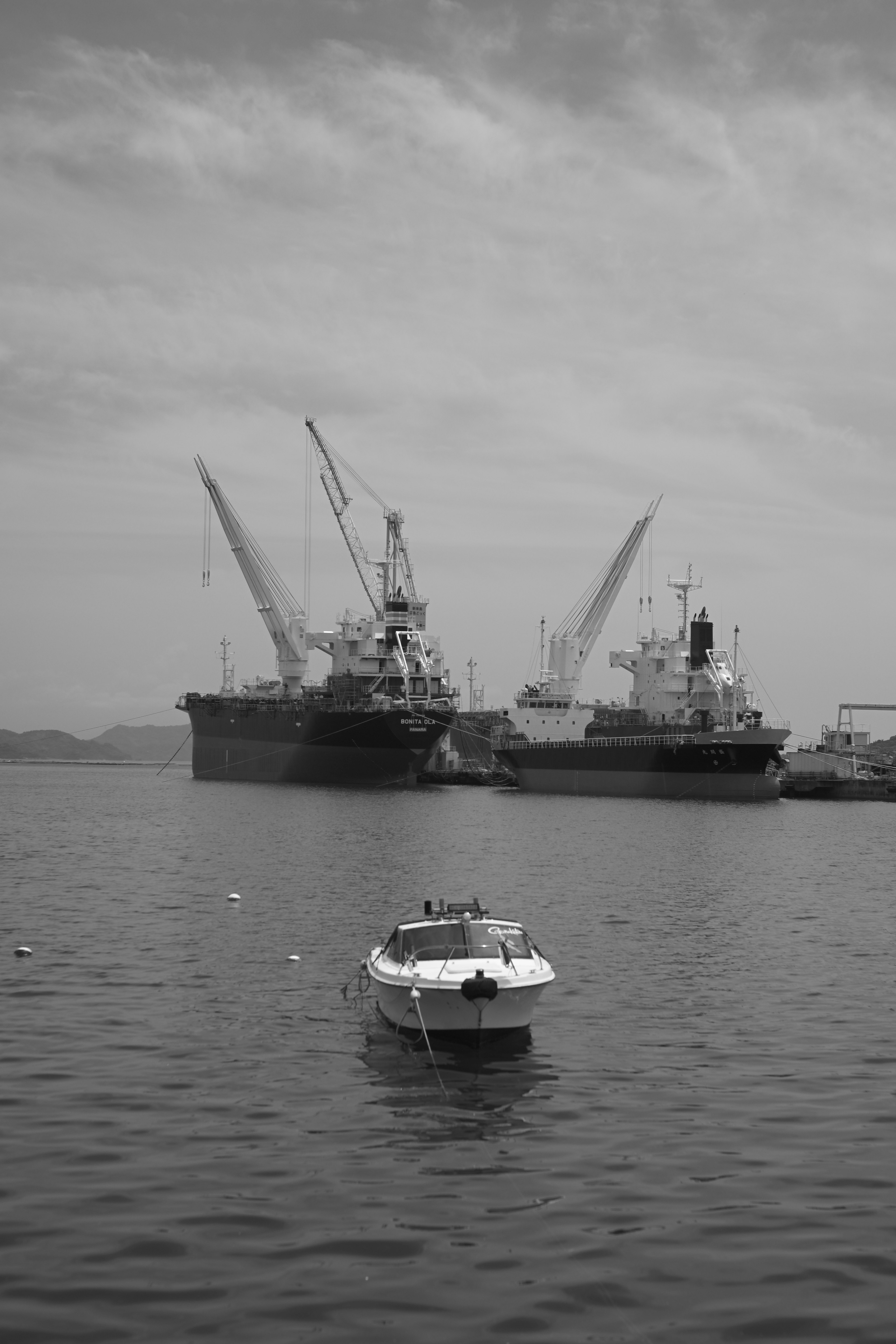 A small boat floating in black and white water with large cargo ships in the background