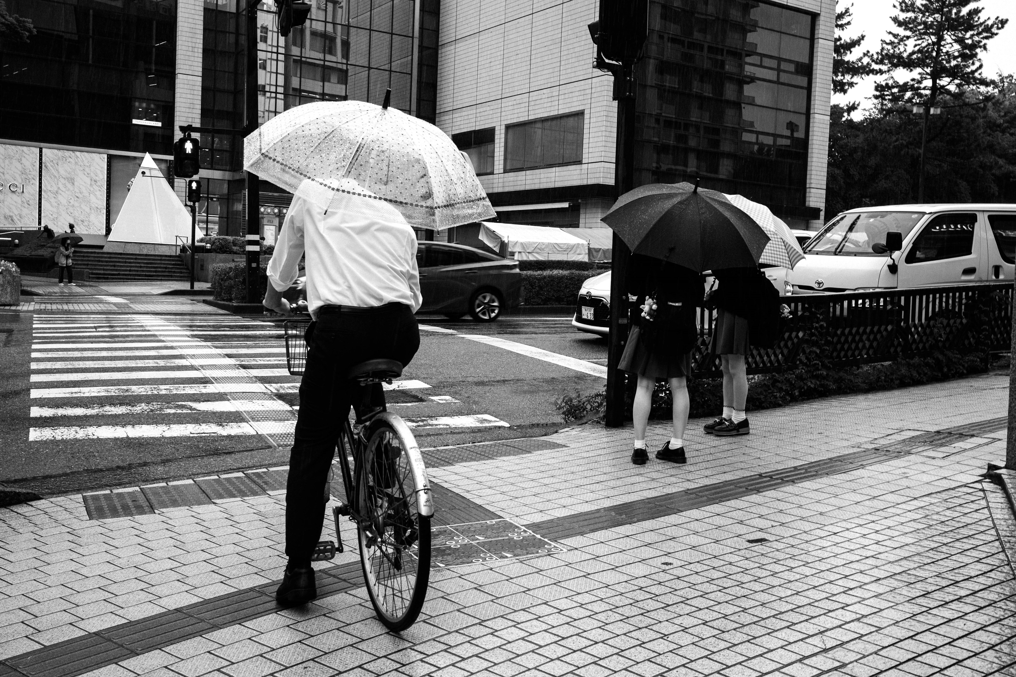 Schwarzweißfoto eines Mannes auf einem Fahrrad mit einem Regenschirm und Fußgängern mit Regenschirmen im Regen