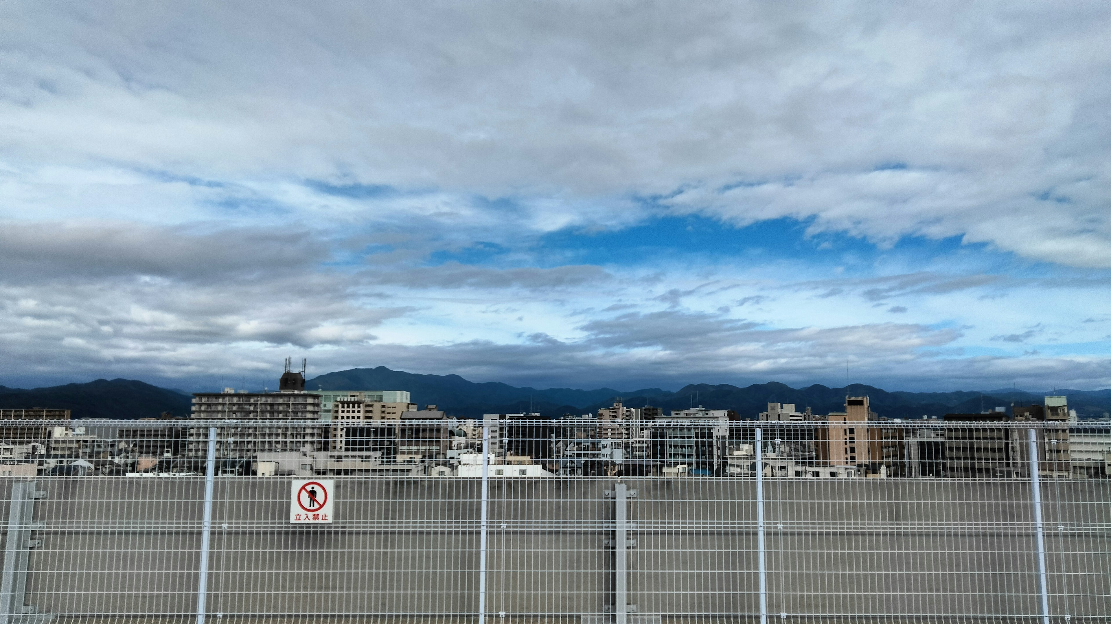 Cityscape with blue sky and clouds featuring a fence in the foreground and distant mountains