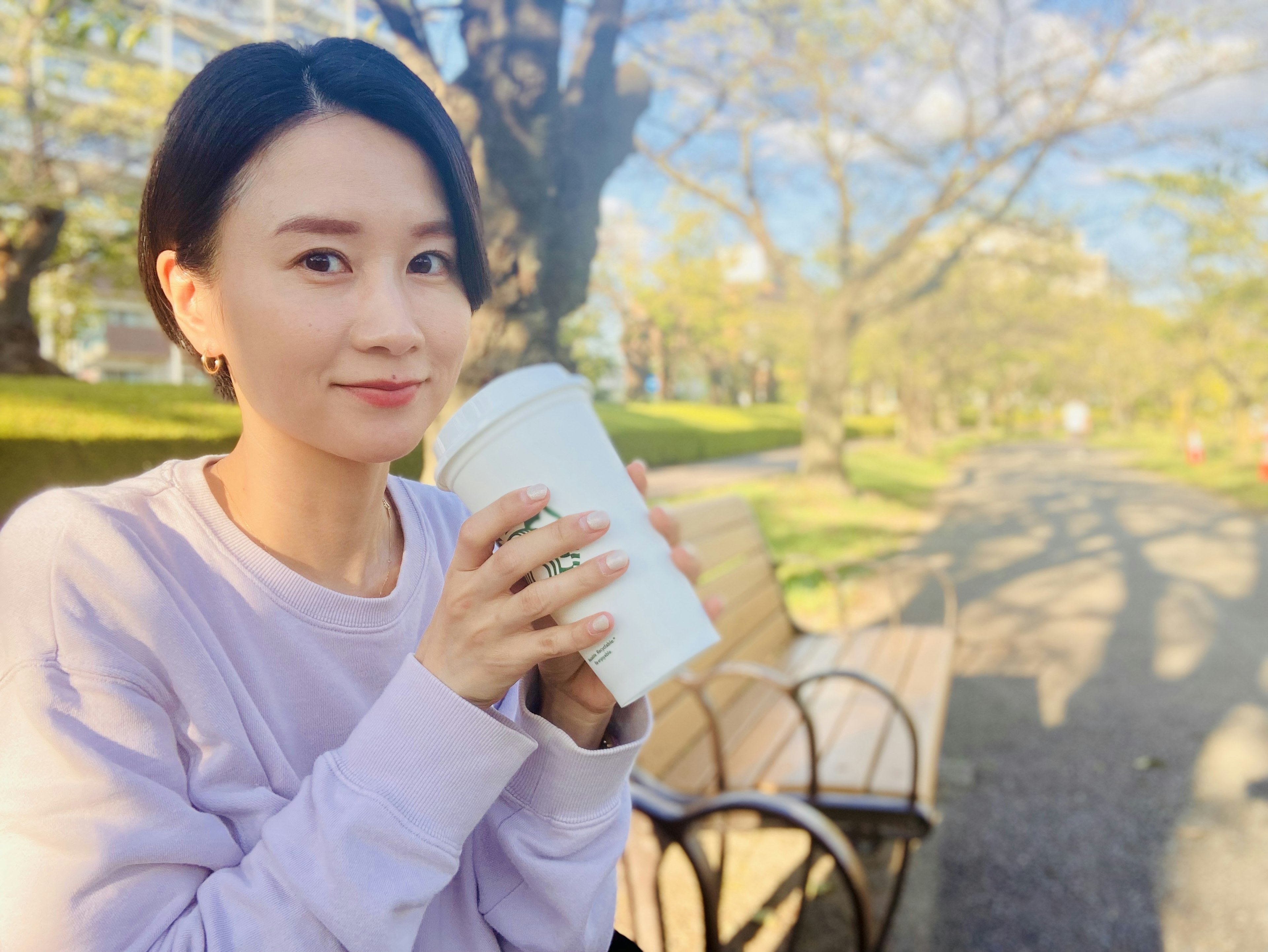 Une femme assise sur un banc de parc tenant une tasse blanche et souriant