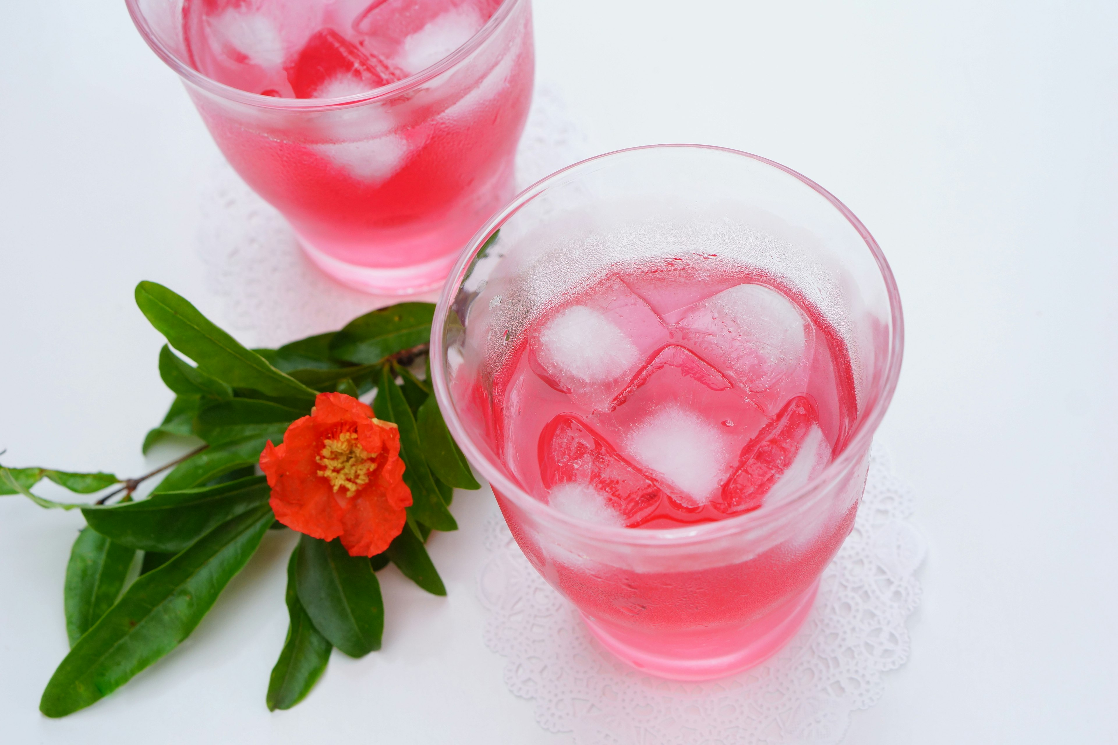 Dos vasos de bebida rosa con hielo acompañados de hojas verdes y una flor roja