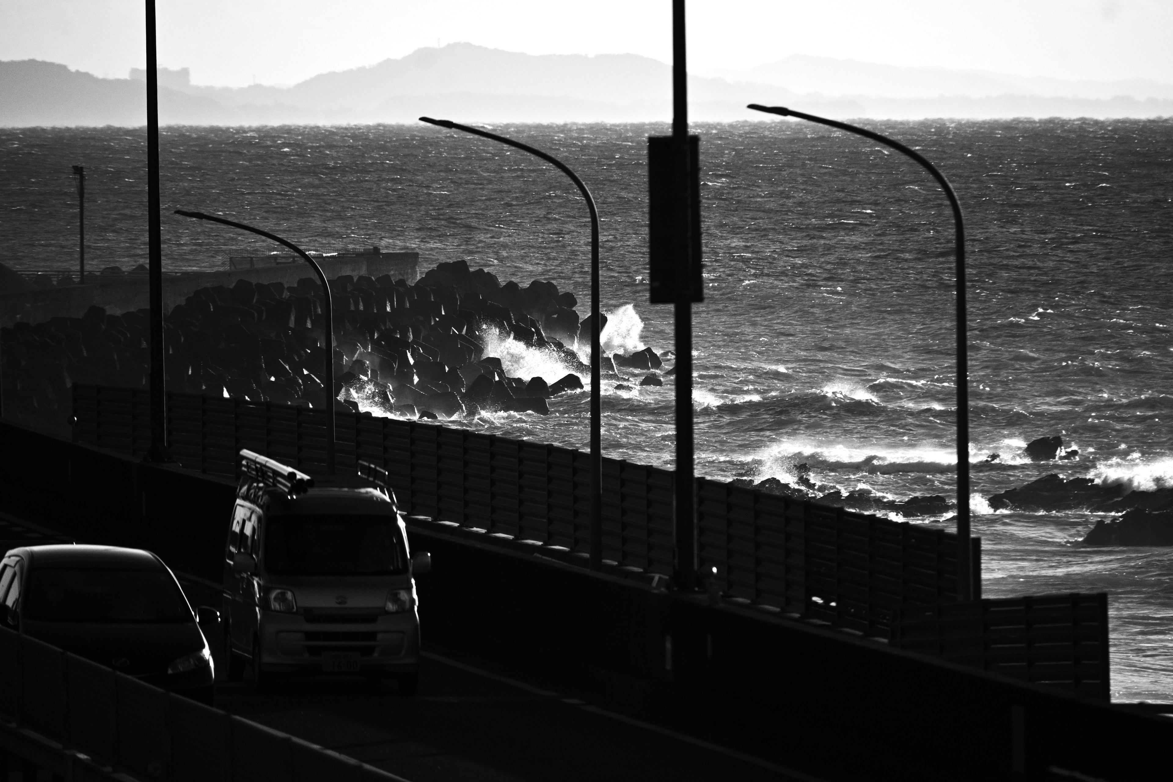 Black and white photo of waves crashing against rocks with cars on a road