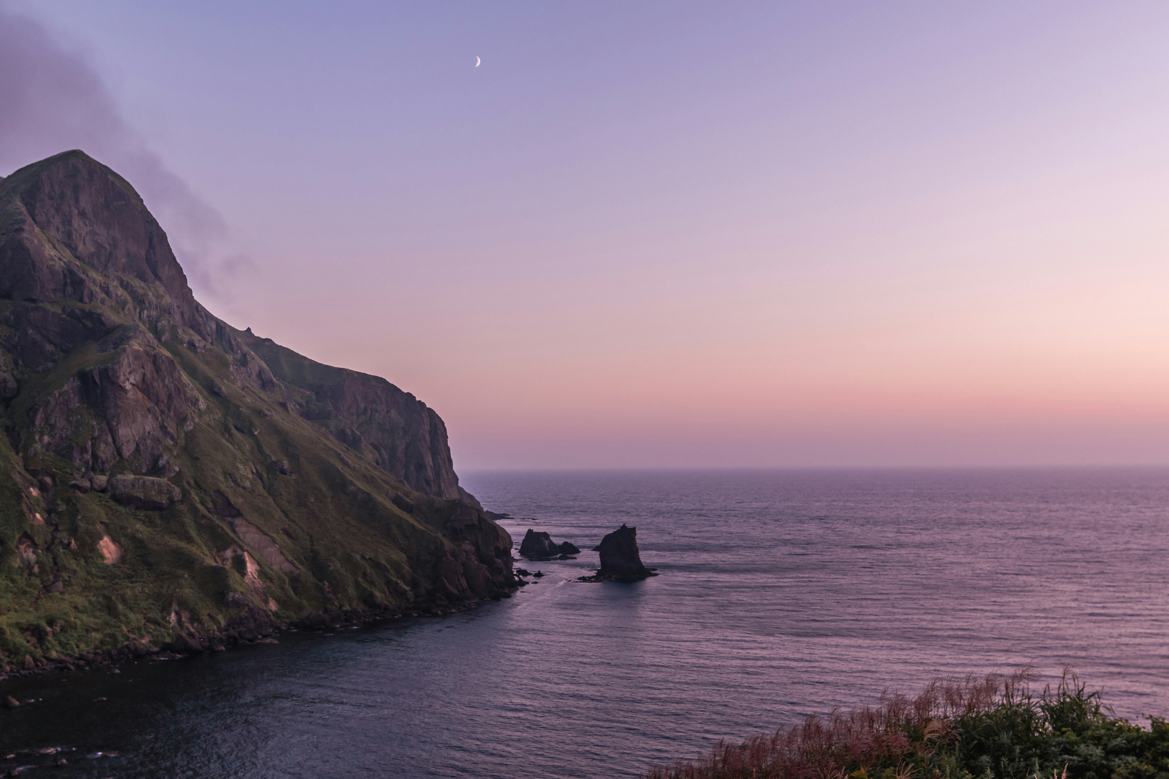 Pemandangan gunung dan laut dengan langit ungu pucat dan bulan terlihat