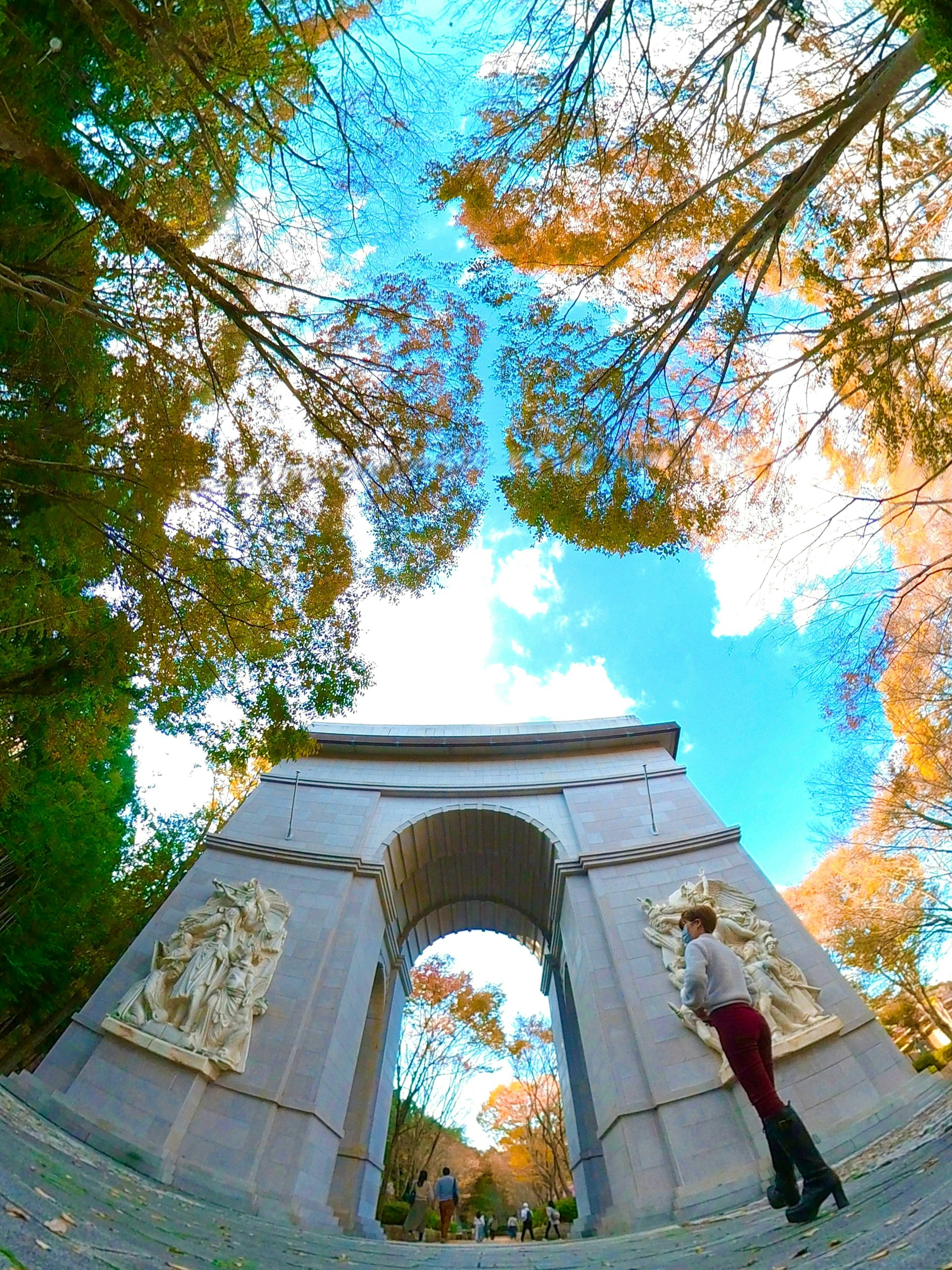 A woman standing under an arch surrounded by colorful trees and a blue sky