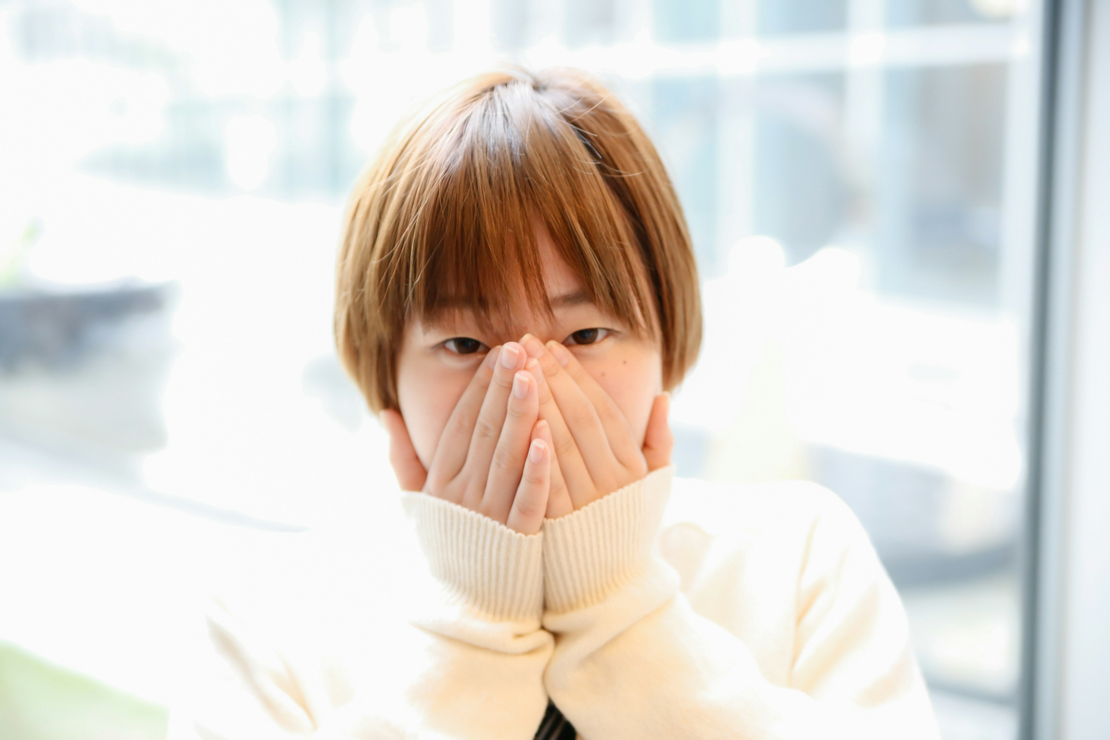 Portrait of a young woman covering her face with her hands in front of a window