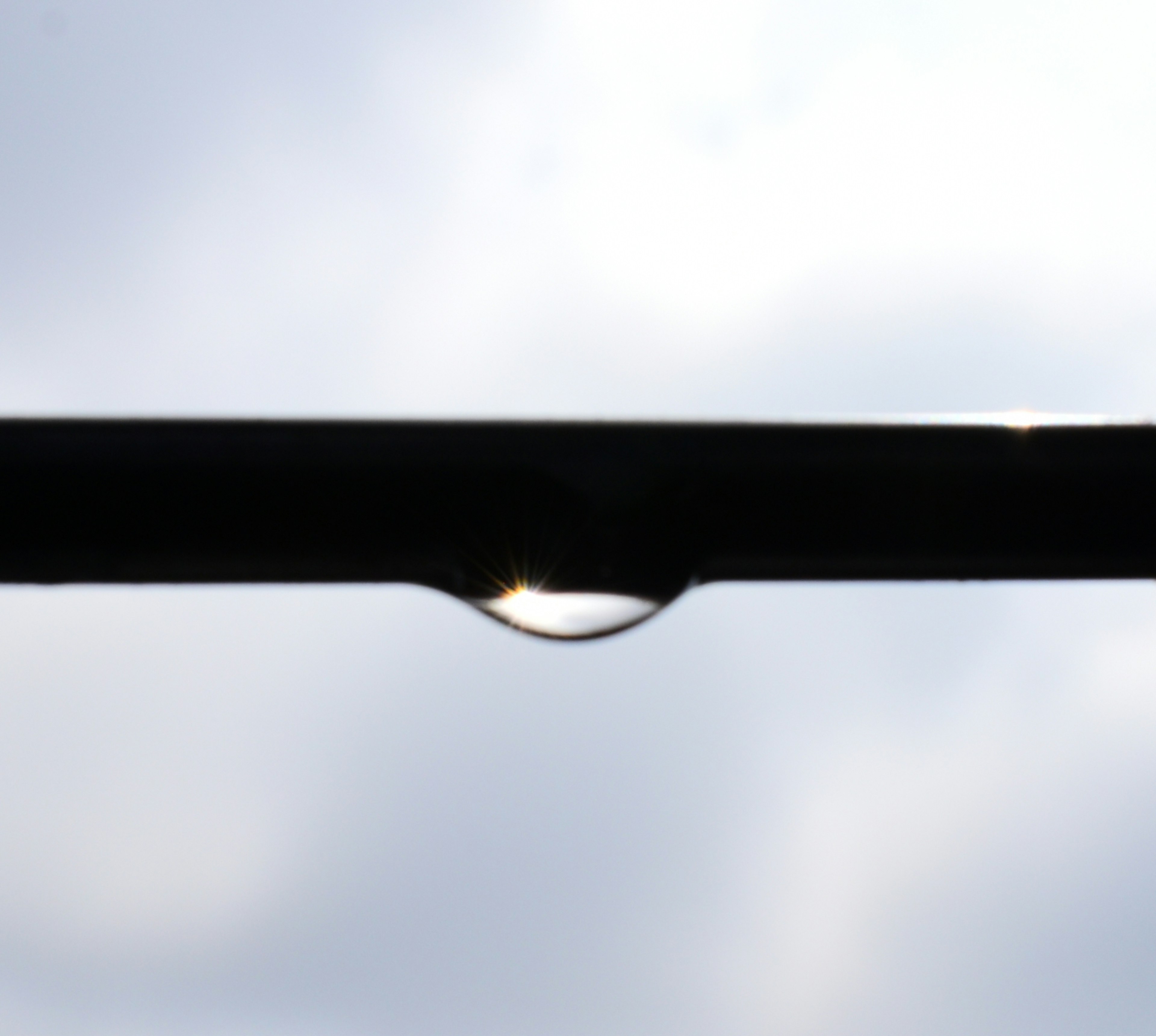 A transparent water droplet hanging from a black rod against a cloudy sky