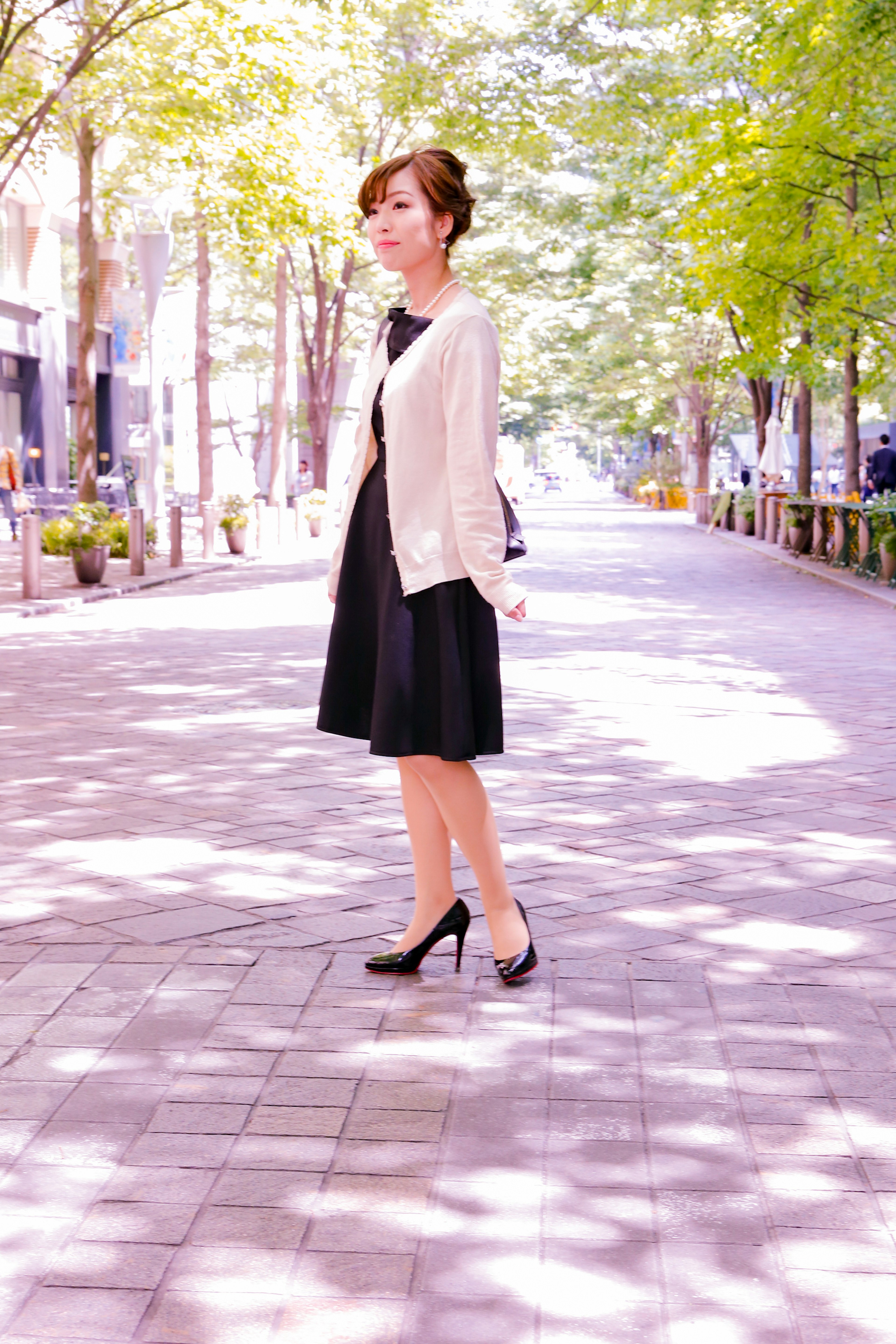 A woman in a black dress and white cardigan standing on a tree-lined street