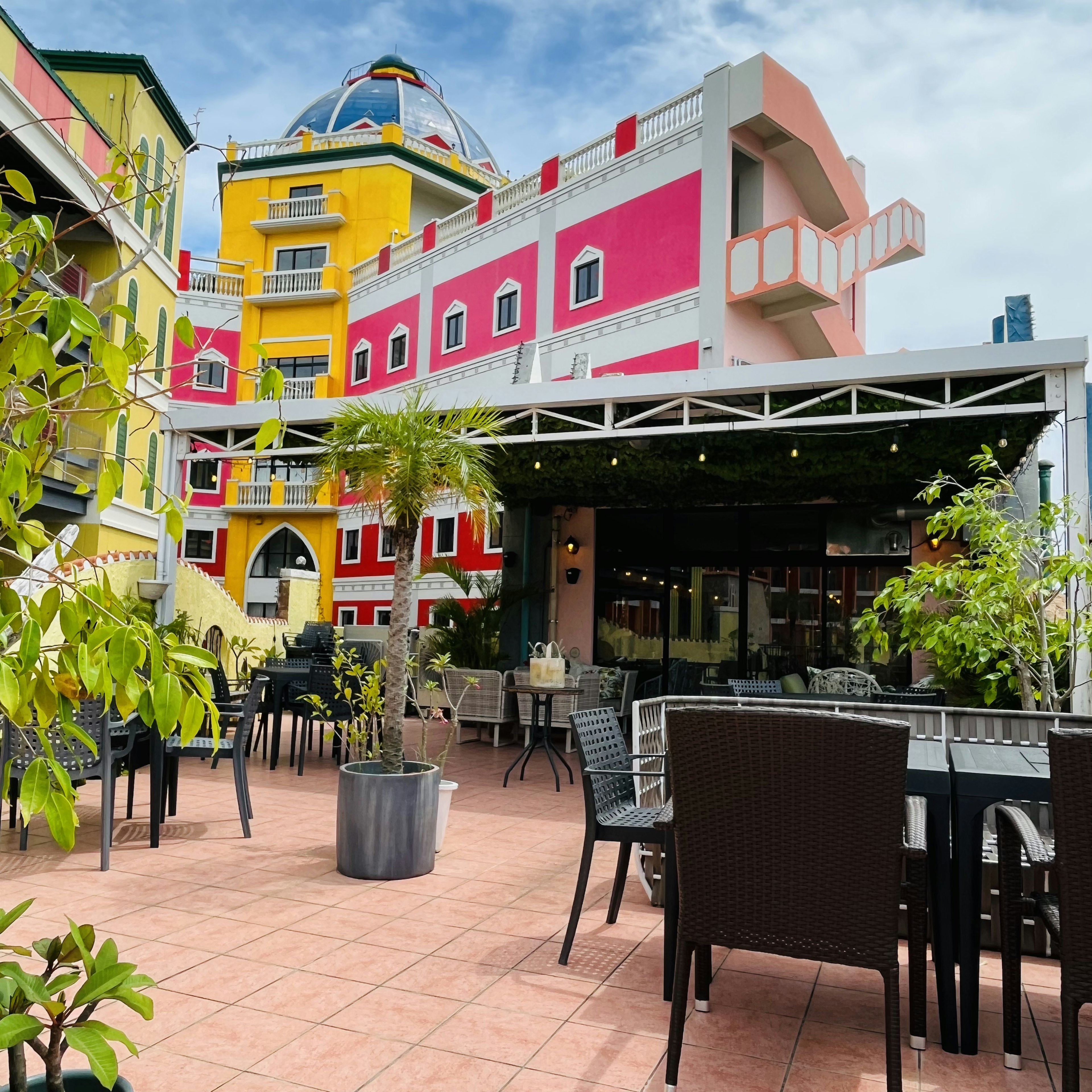 Área de comedor al aire libre rodeada de edificios coloridos con plantas en macetas y mesas