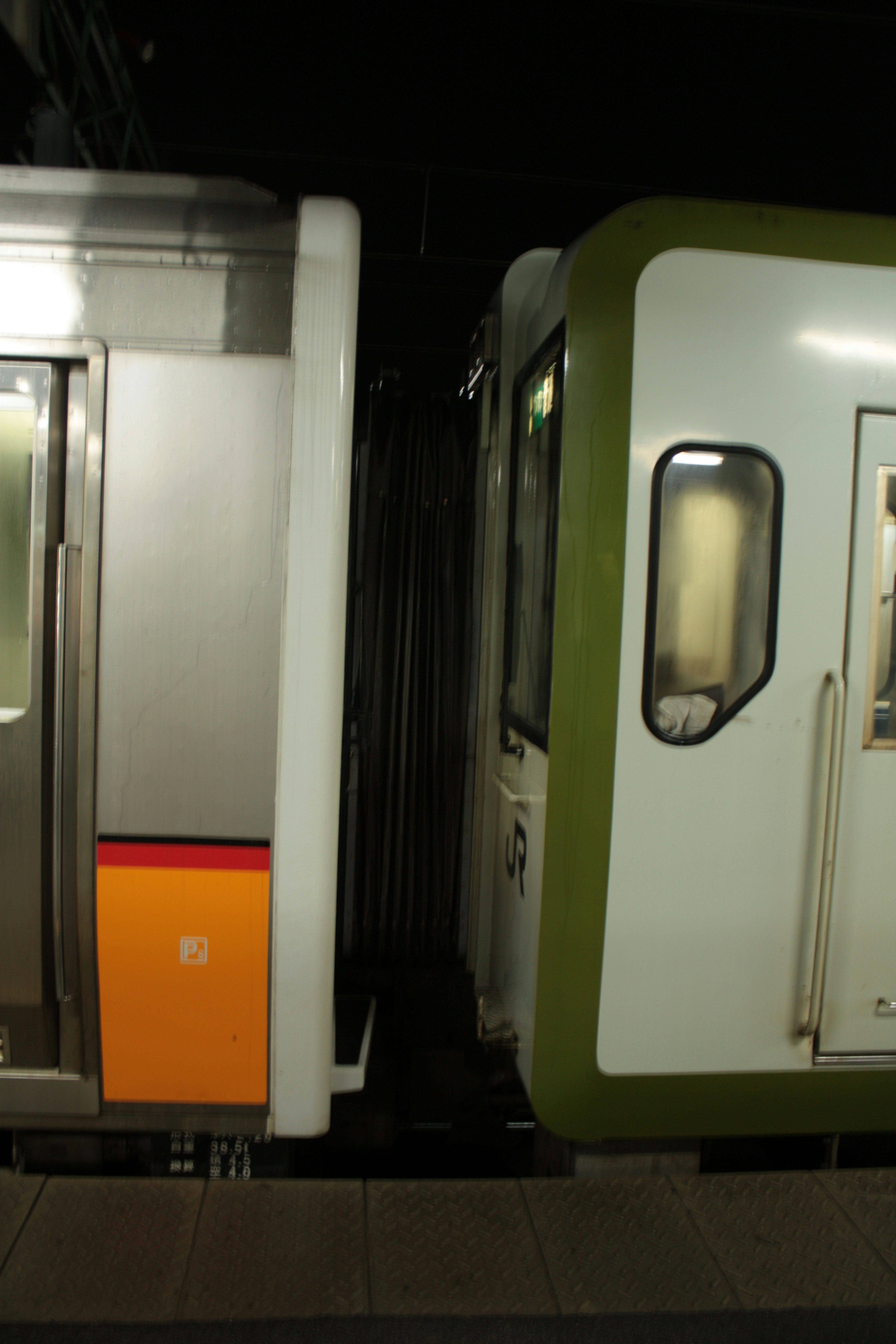 Dos vagones de tren uno plateado con detalles naranjas y otro verde y blanco en una estación poco iluminada