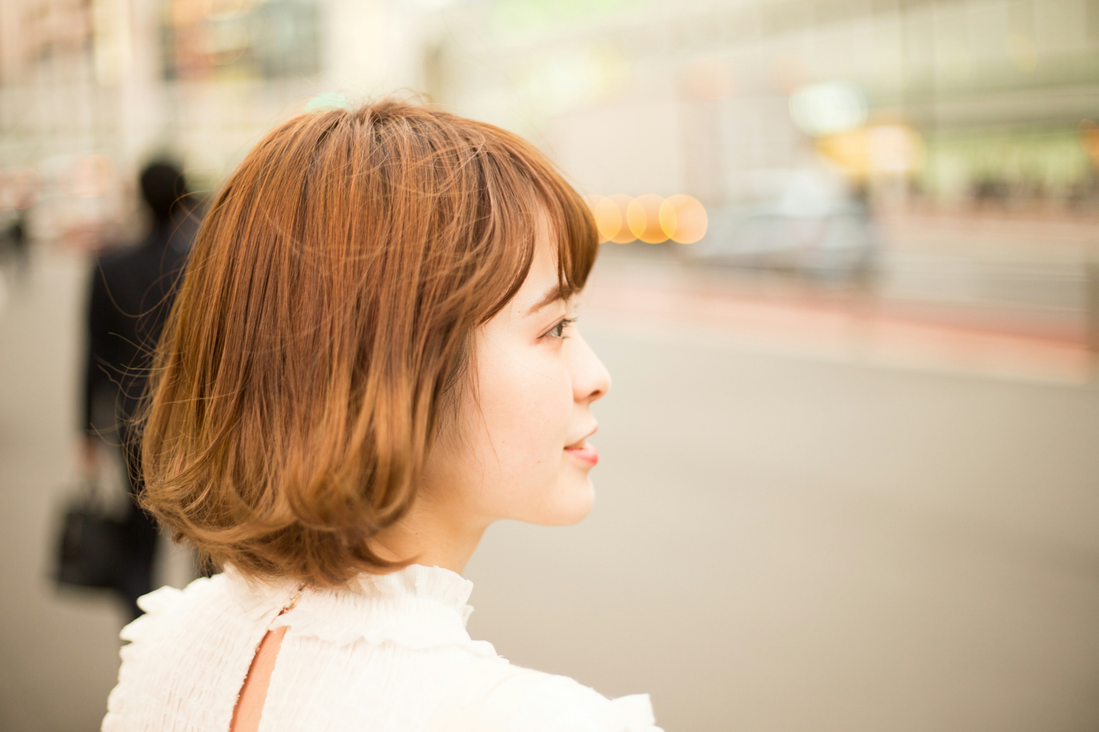 Profile of a woman looking at the street with short brown hair and a white outfit blurred pedestrians and cars in the background