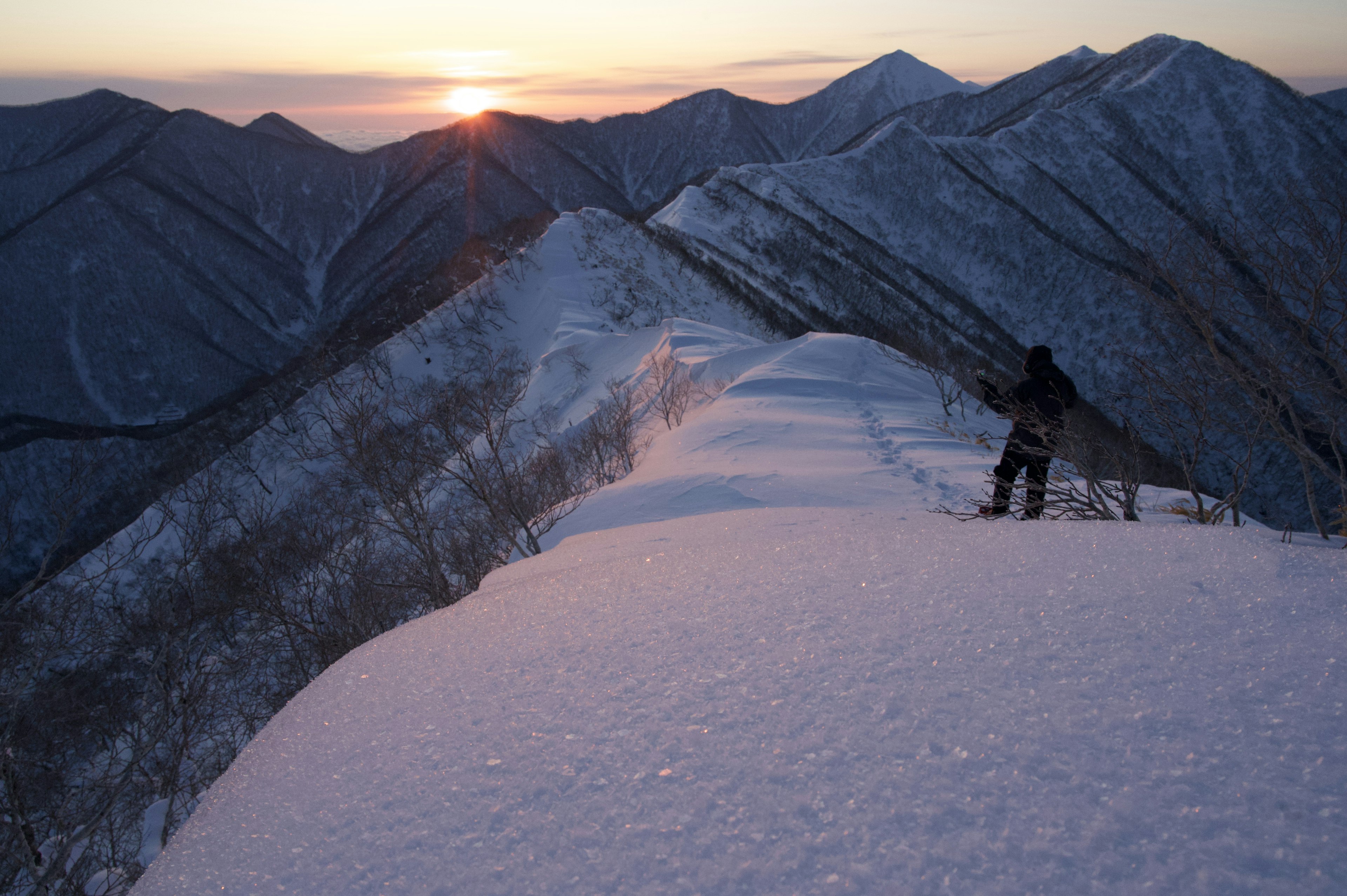 雪に覆われた山の稜線と夕日をバックに立つ登山者