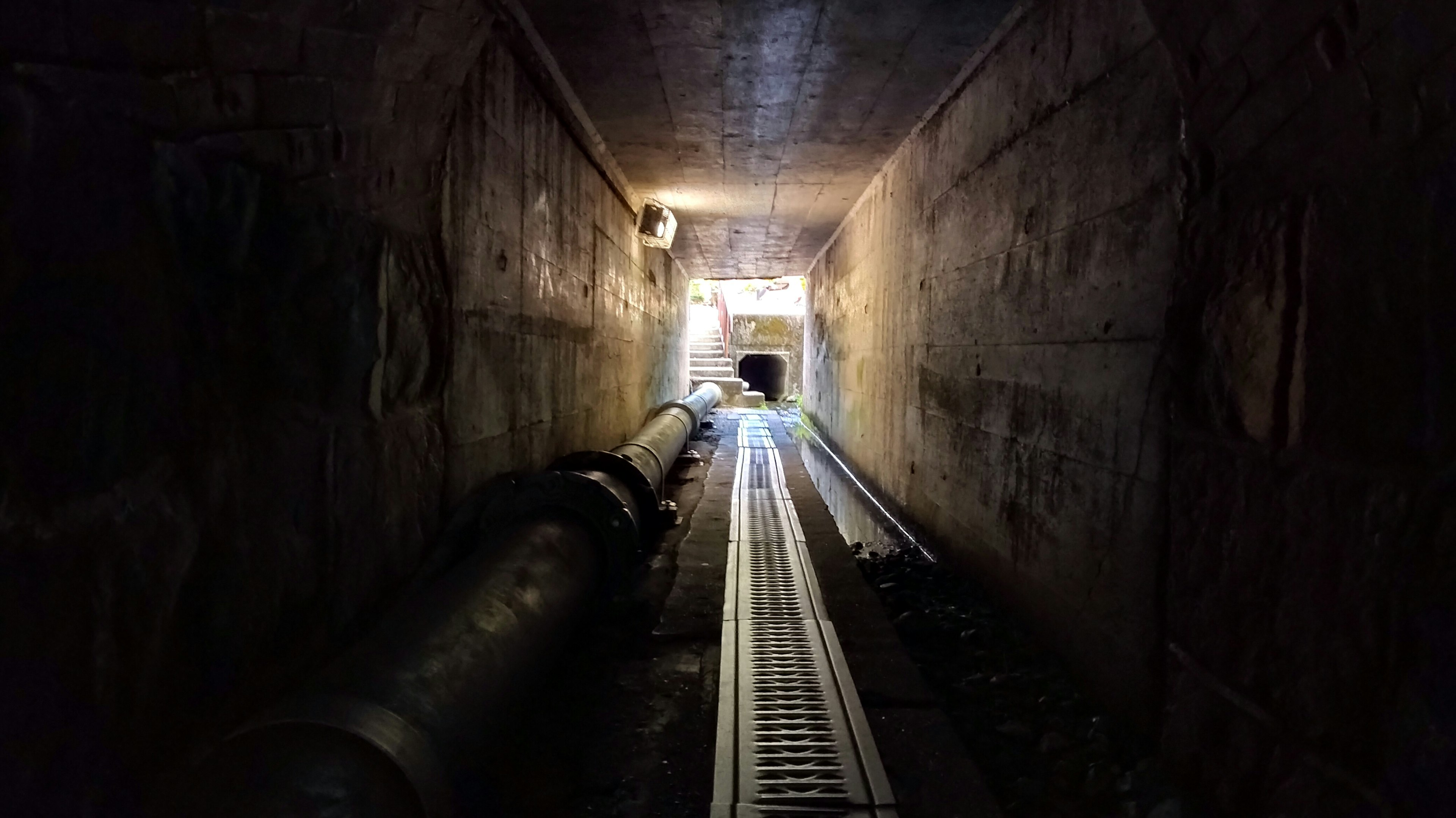 Dimly lit tunnel with pipes and tracks leading towards a bright exit