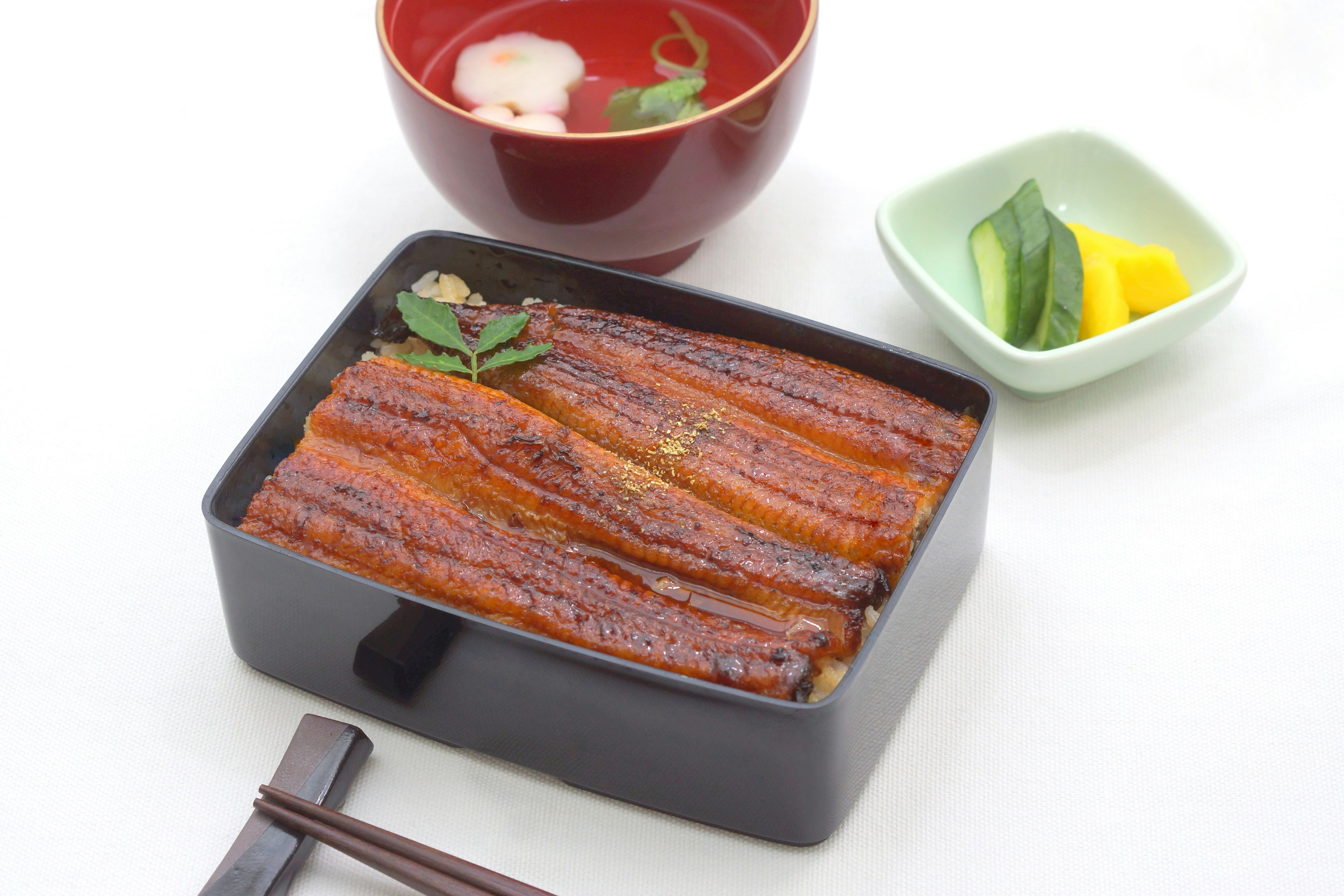 Anguille grillée servie dans une boîte noire avec de la soupe miso dans un bol rouge et des cornichons dans un plat vert