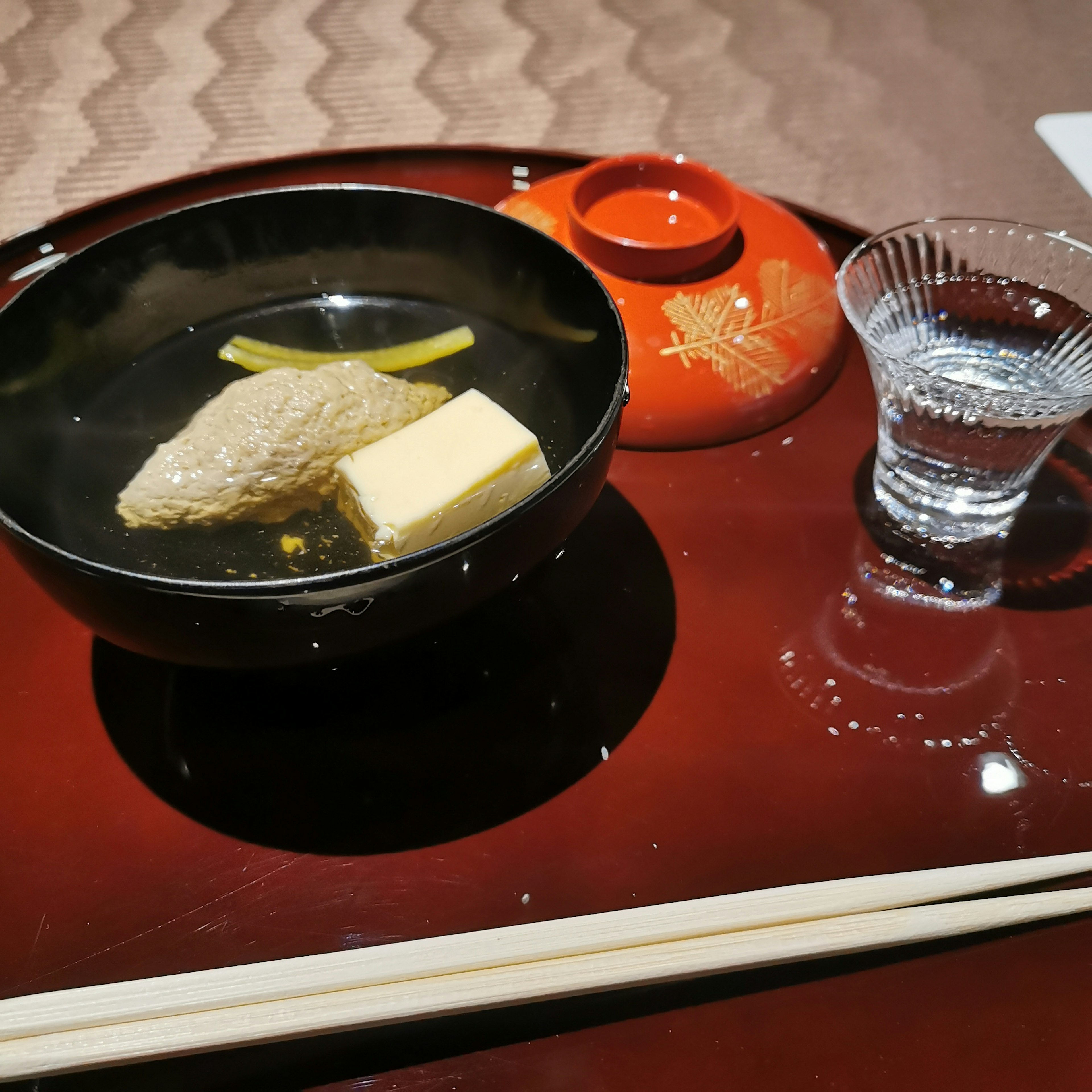 Un bol noir avec un plat de tofu et de poisson accompagné d'un verre de saké et d'un contenant rouge