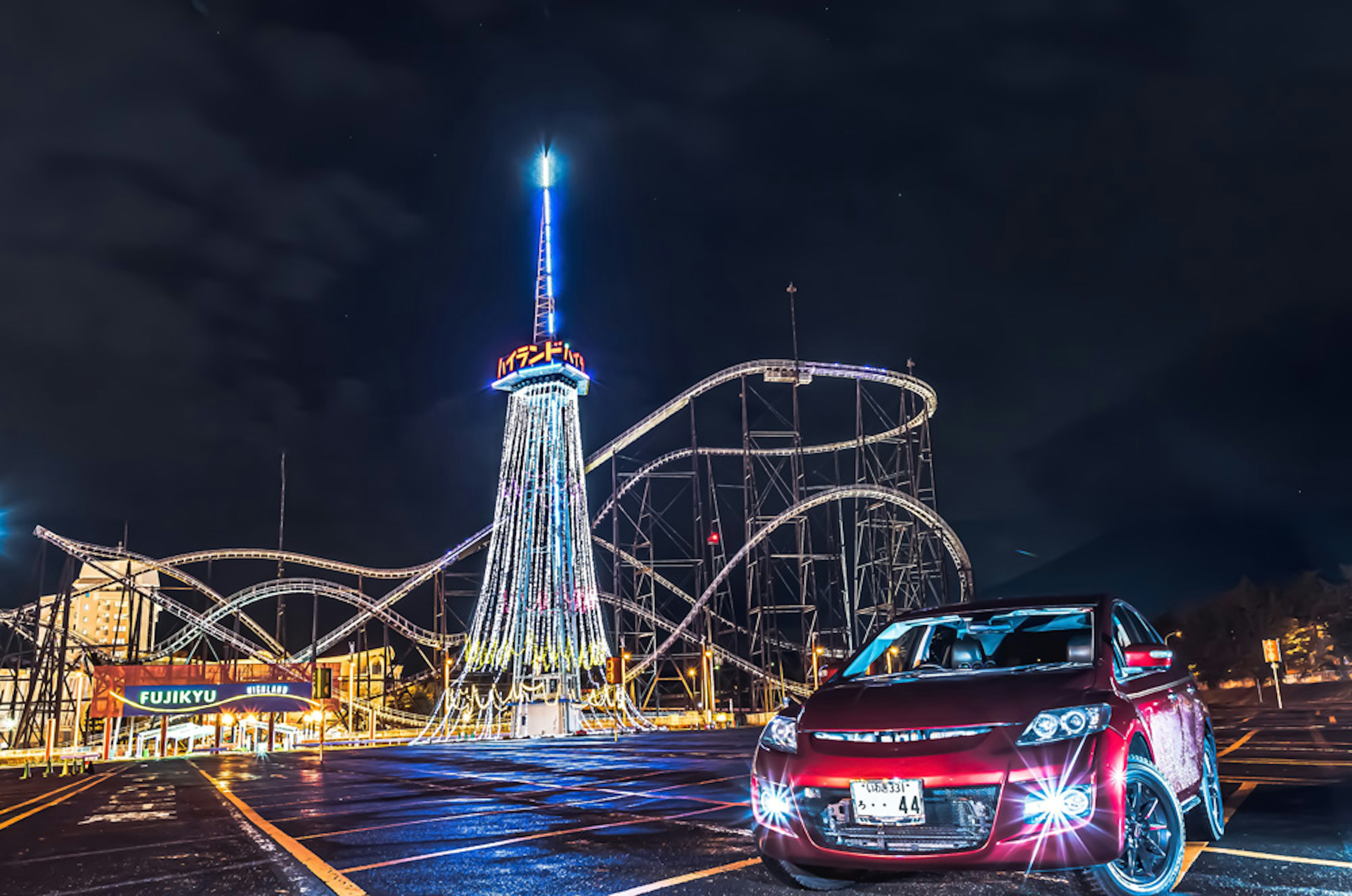 Mobil merah diparkir di depan taman hiburan yang terang dan roller coaster di malam hari