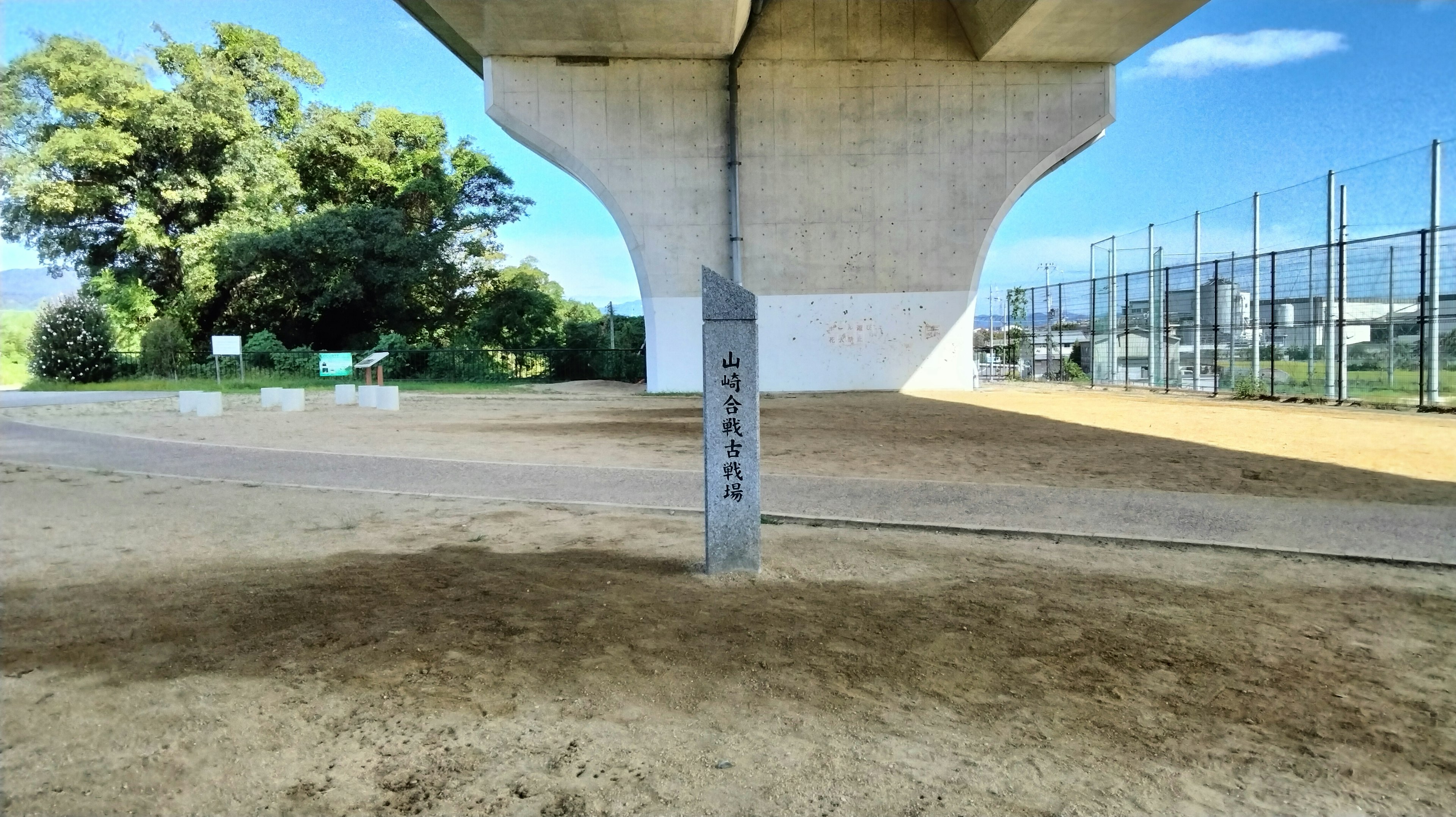 A sandy park area beneath a large concrete bridge pillar with a baseball field in the background