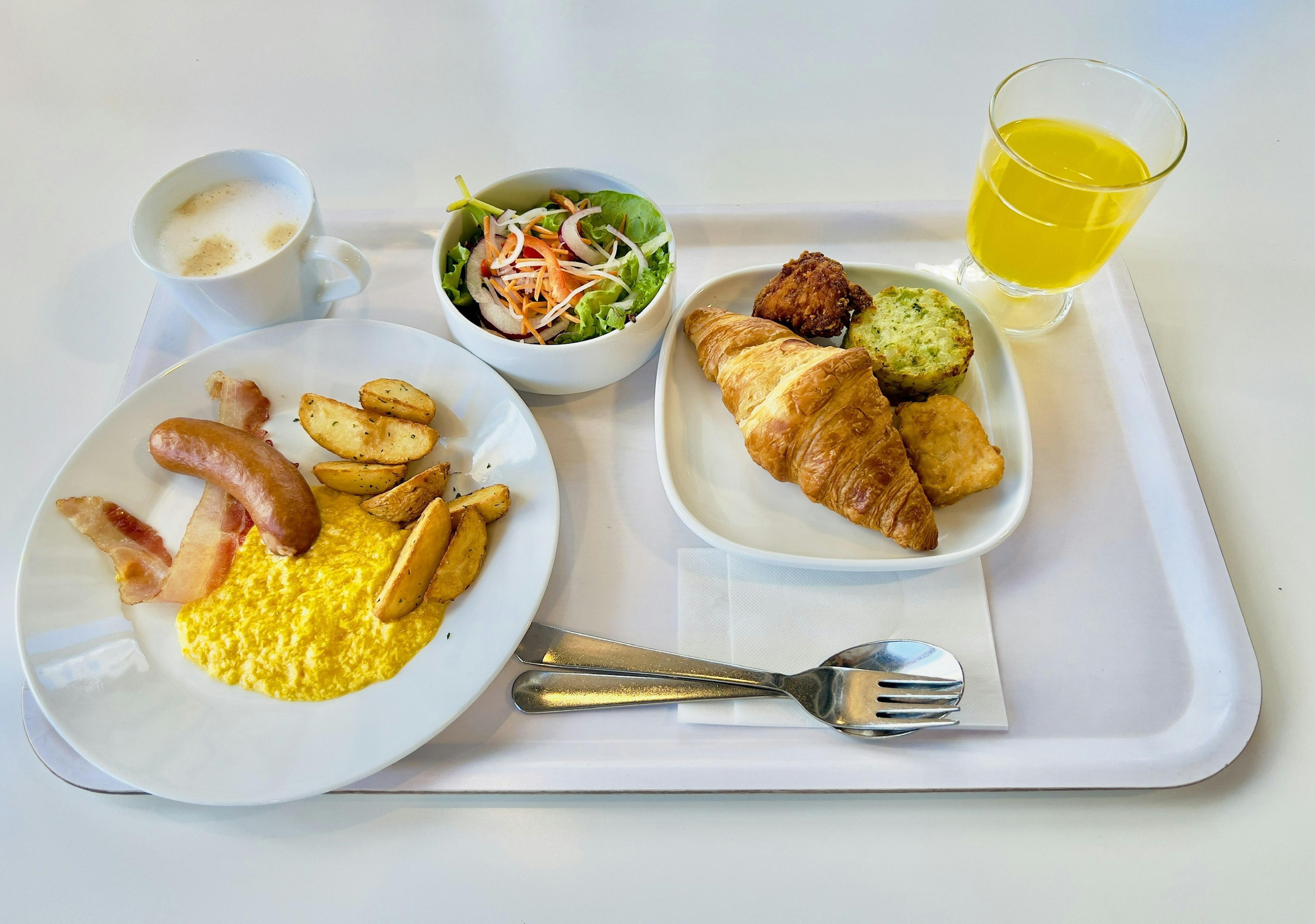 Bandeja de desayuno con huevos revueltos, salchicha, papas fritas, ensalada, croissant, jugo de naranja