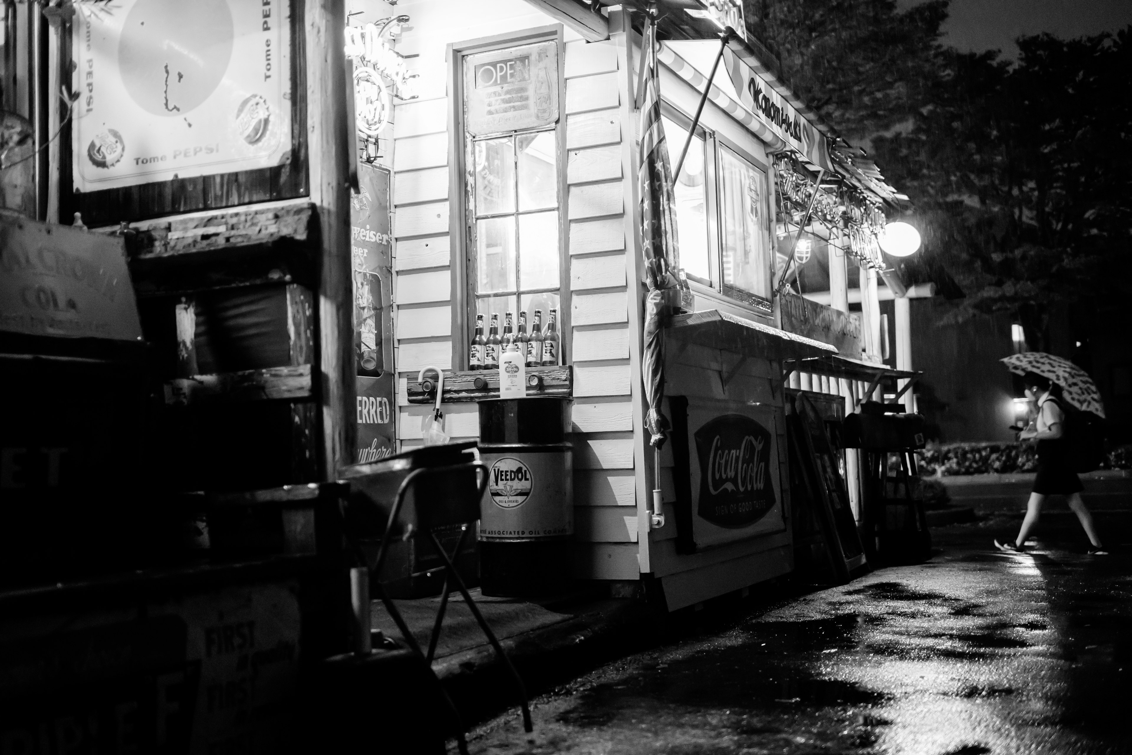 Schwarzweißfoto eines Straßenimbisses bei Nacht mit glänzendem Gehweg und warmem Licht