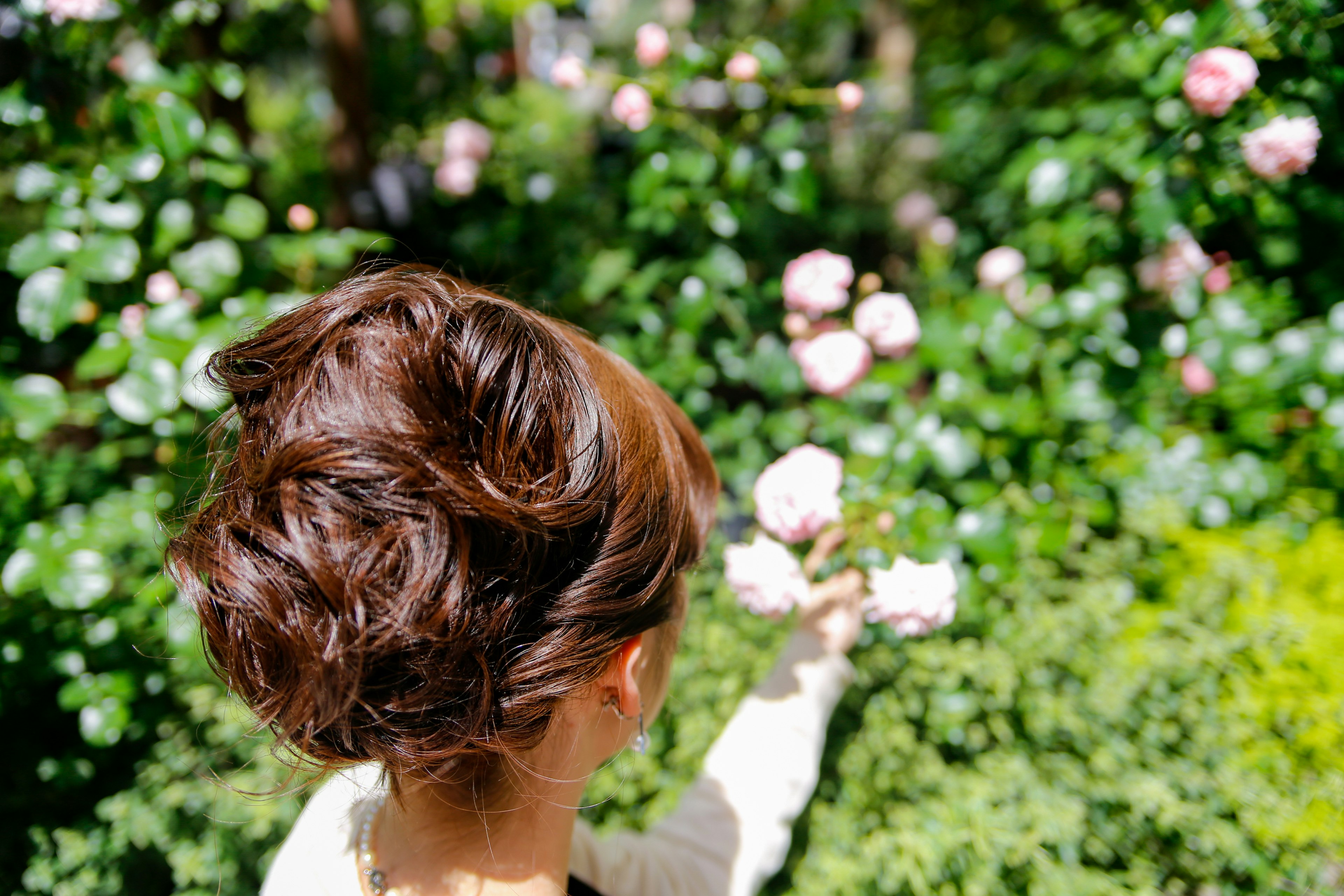 Una mujer sosteniendo una flor mientras está de espaldas con un fondo verde exuberante y flores rosas en flor