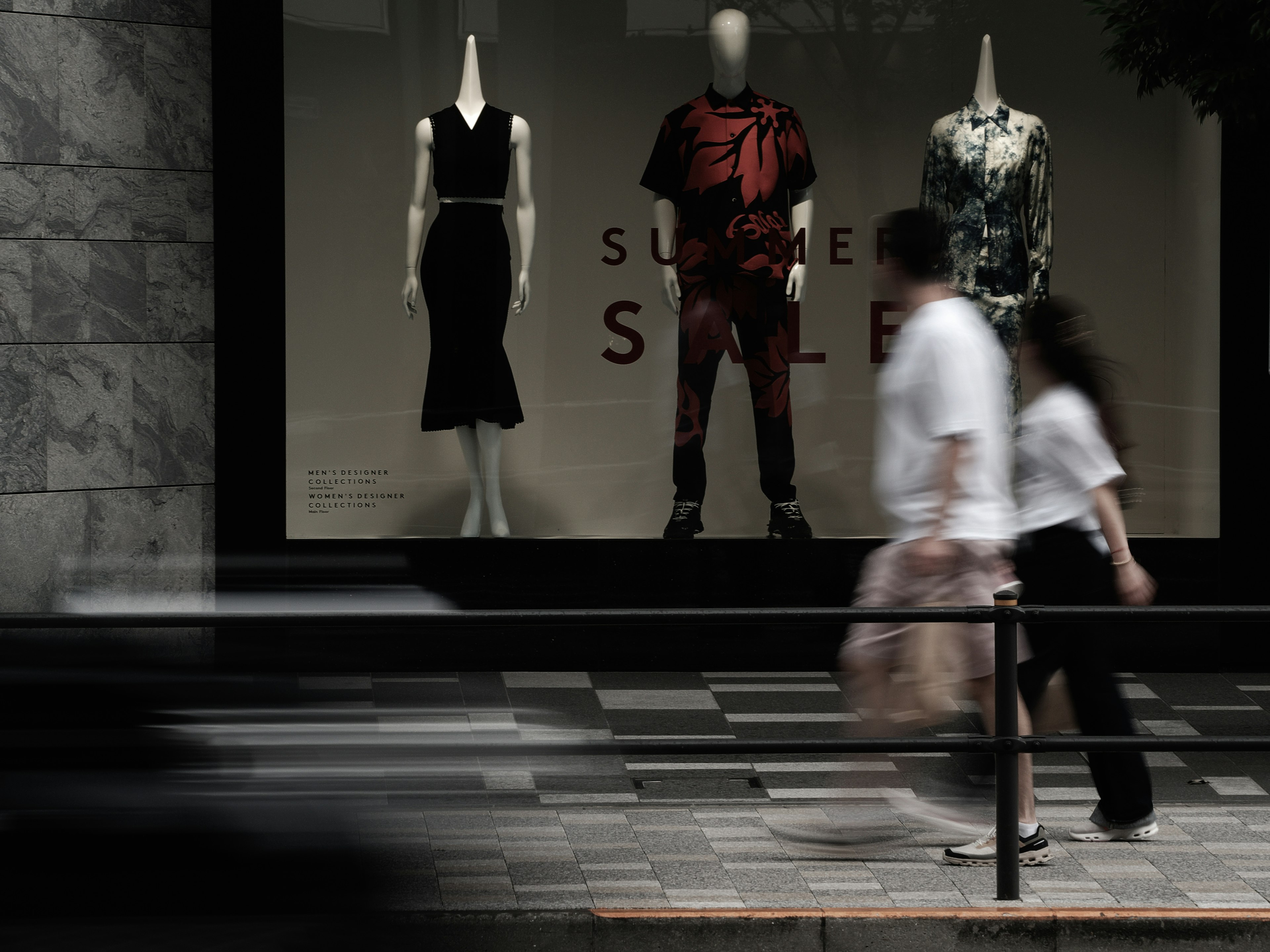 Street scene featuring mannequins in a shop window with a summer sale sign and pedestrians