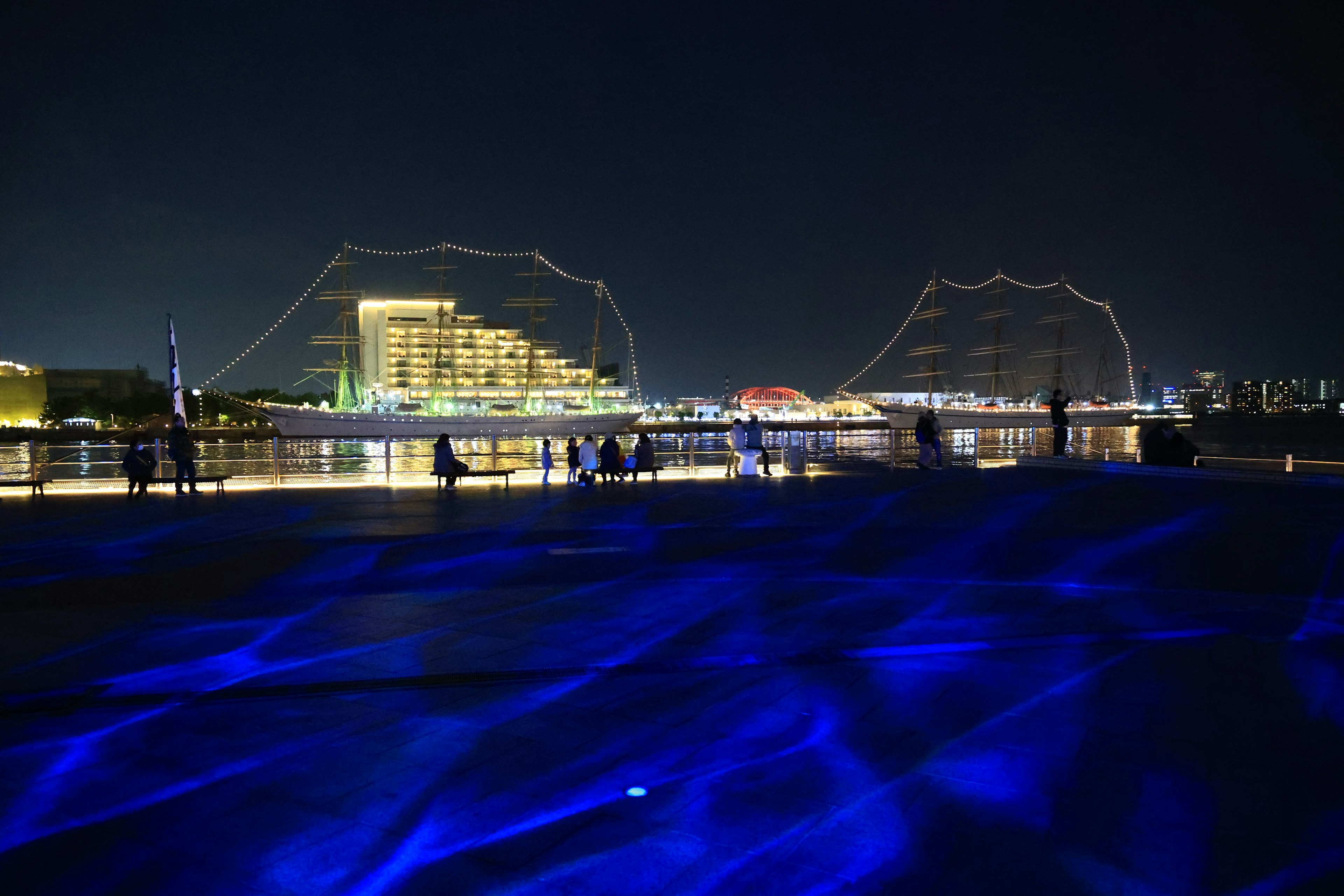 Vista nocturna de un paseo marítimo con reflejos azules y horizonte urbano