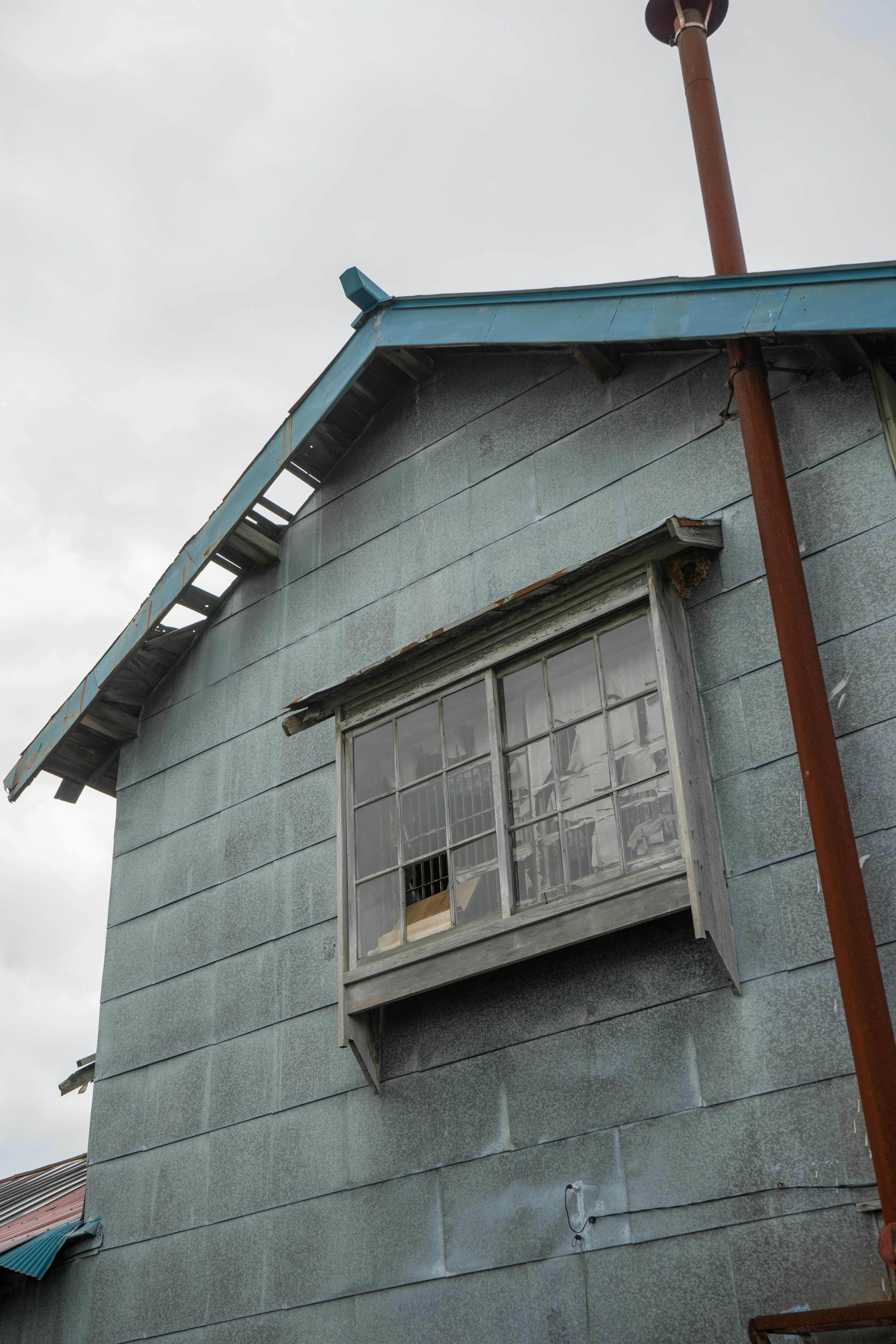 Seitenansicht eines alten Hauses mit einem blauen Dach, das ein Fenster und blau-graue Wände mit einem rostigen Rohr zeigt