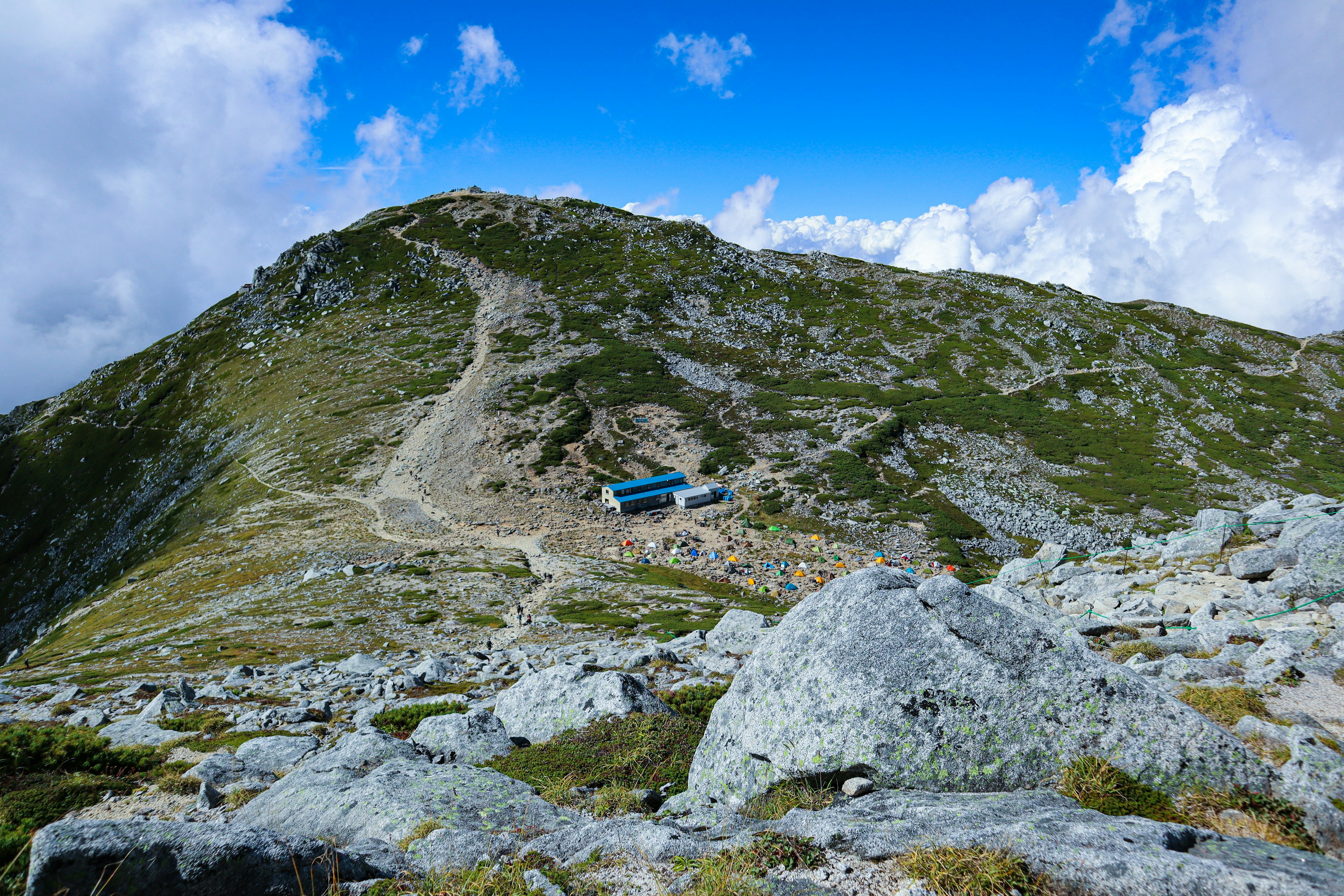 Mountain landscape with a blue cabin, rocky path, green grass, blue sky and white clouds