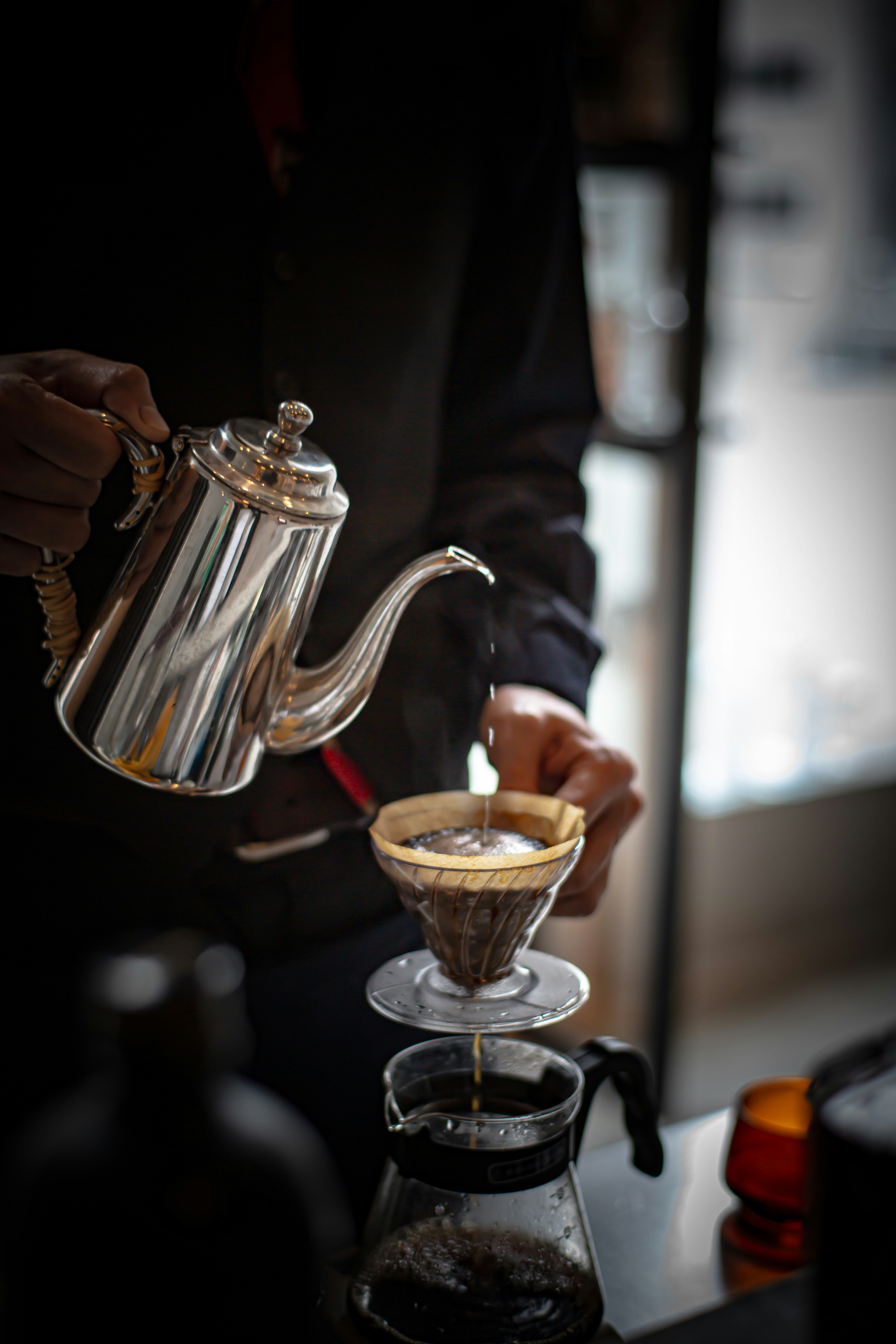 A close-up of a hand pouring coffee from a silver teapot into a coffee filter