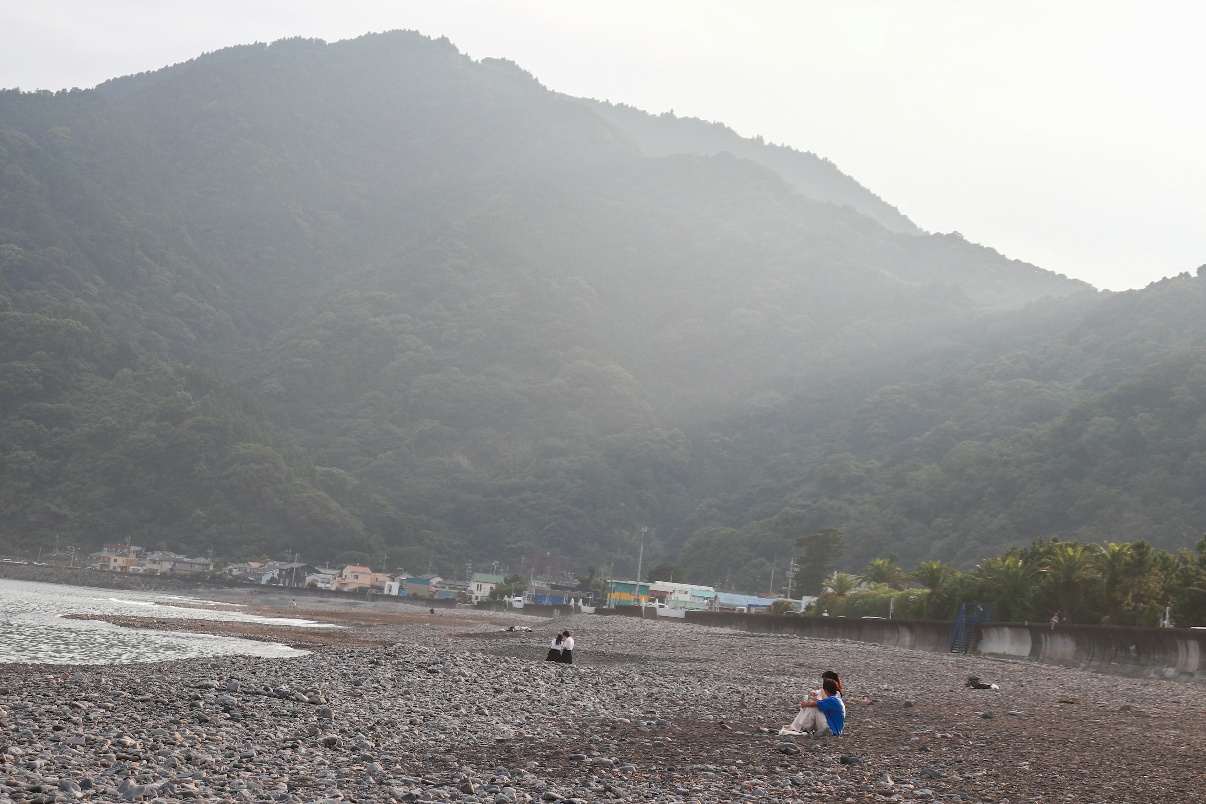 Orang-orang duduk di pantai berbatu dengan latar belakang gunung berkabut