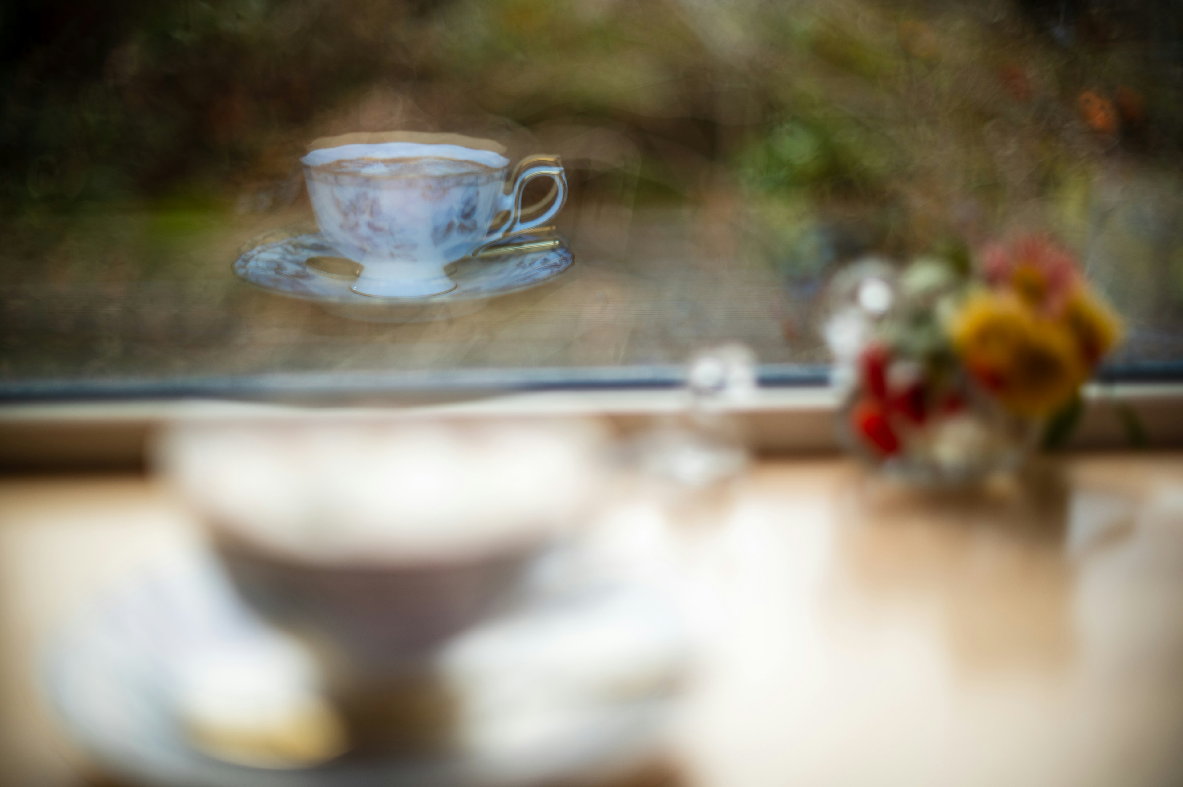 A blurred image featuring a cup seen through a window with flowers on a table