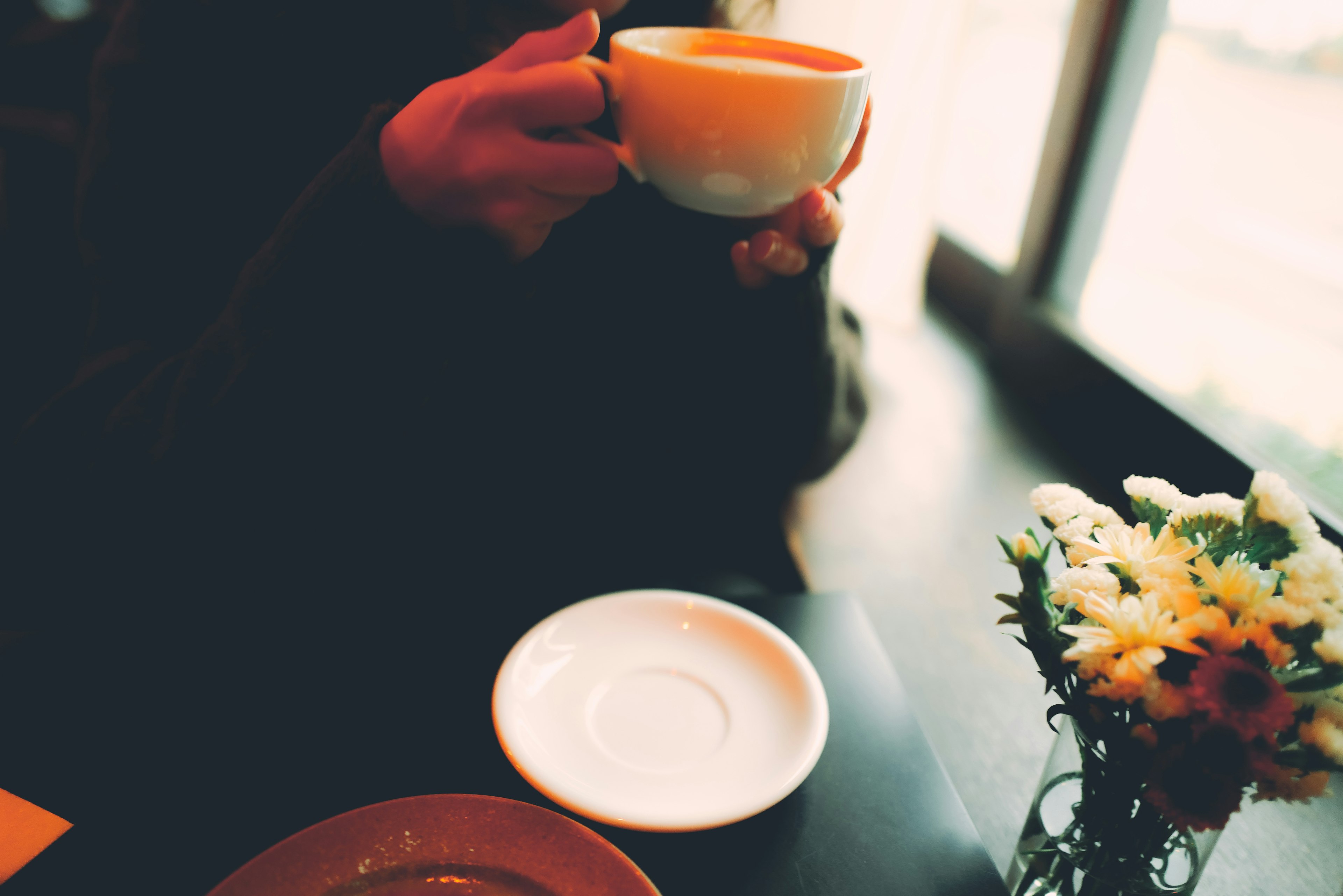 A hand holding a cup at a cafe table with a floral arrangement