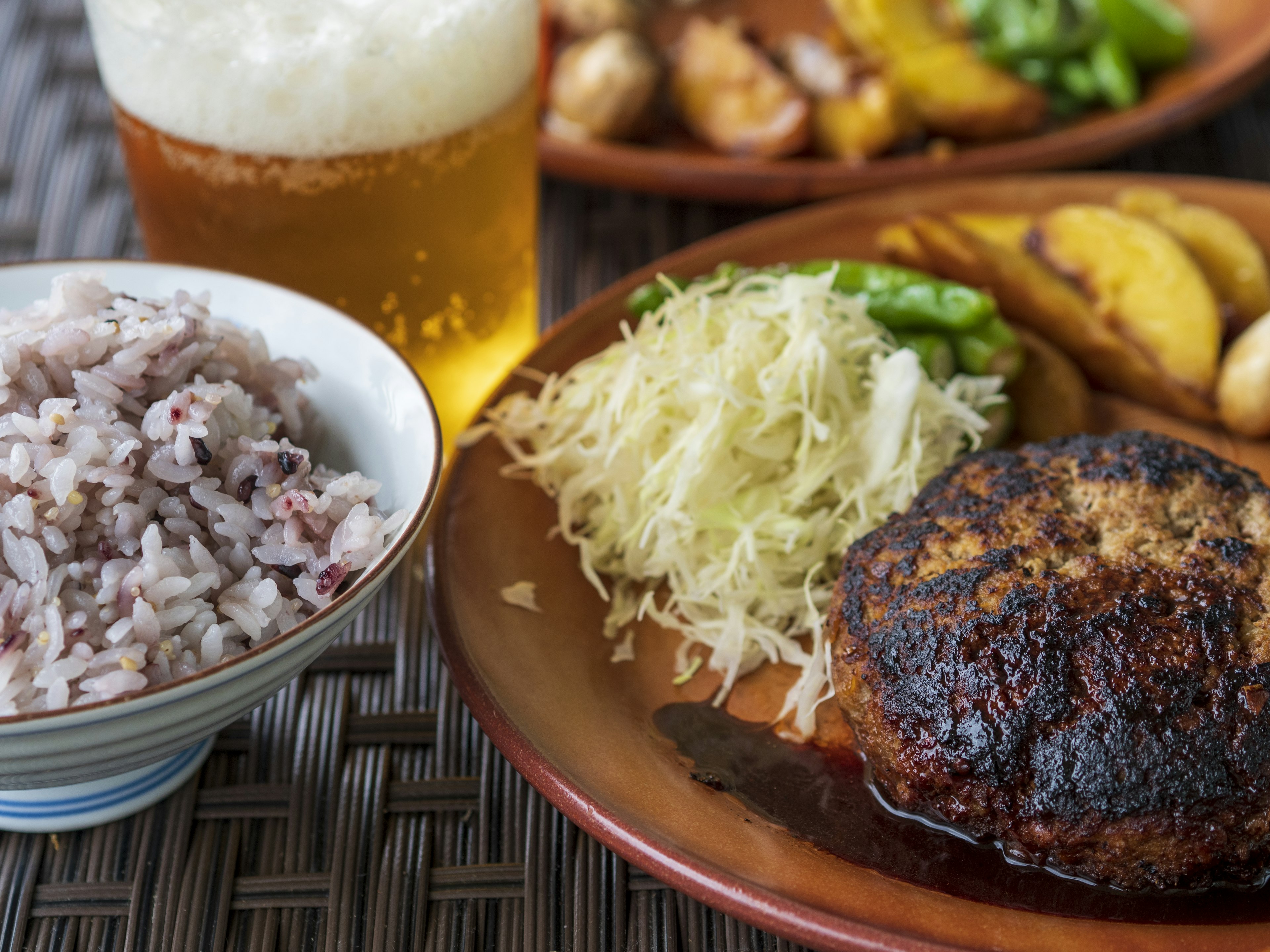 A plate featuring a grilled hamburger steak, shredded cabbage, green peppers, brown rice, and a glass of beer