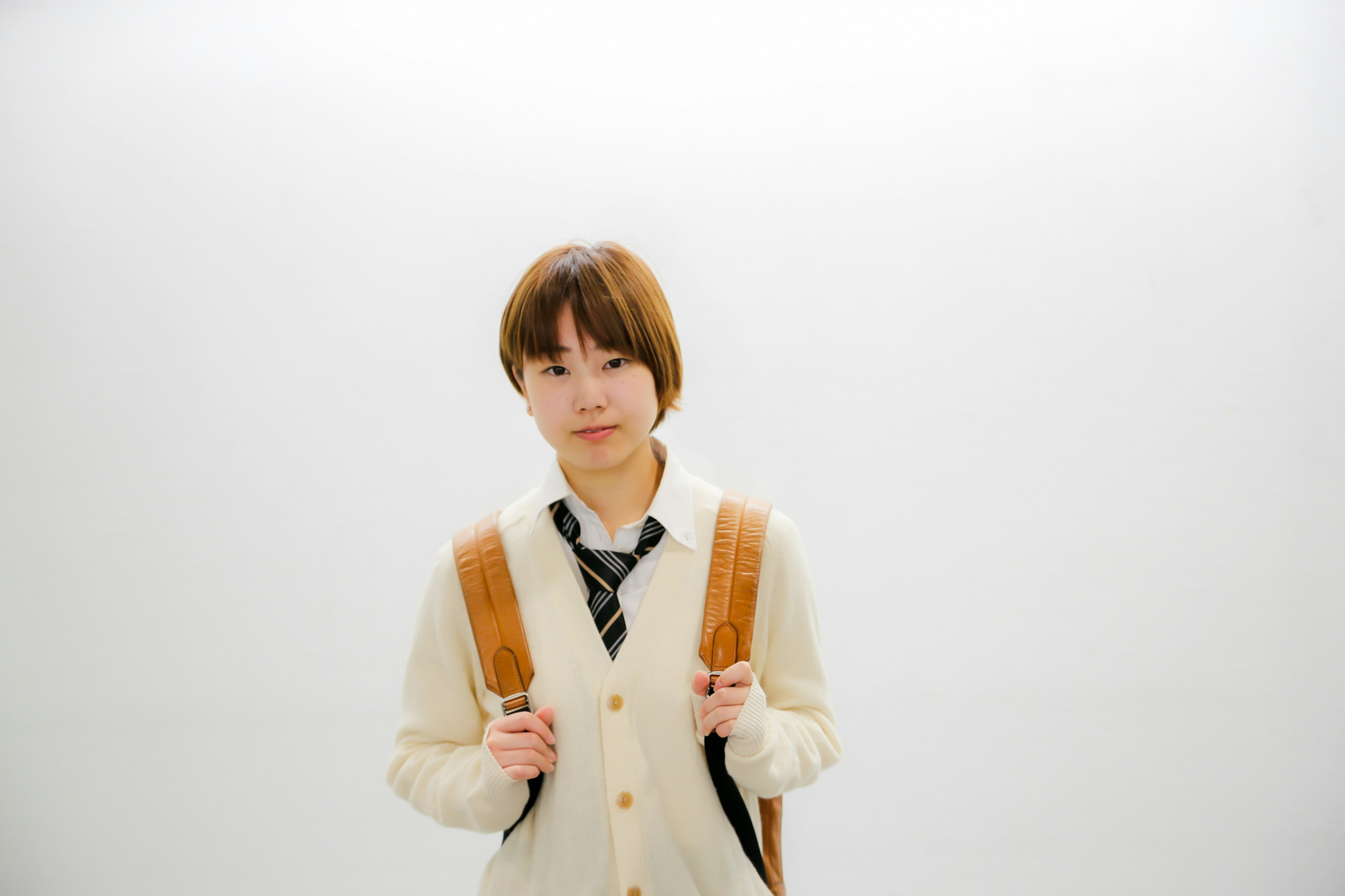 Young woman in a beige sweater and tie standing in front of a white background with a backpack