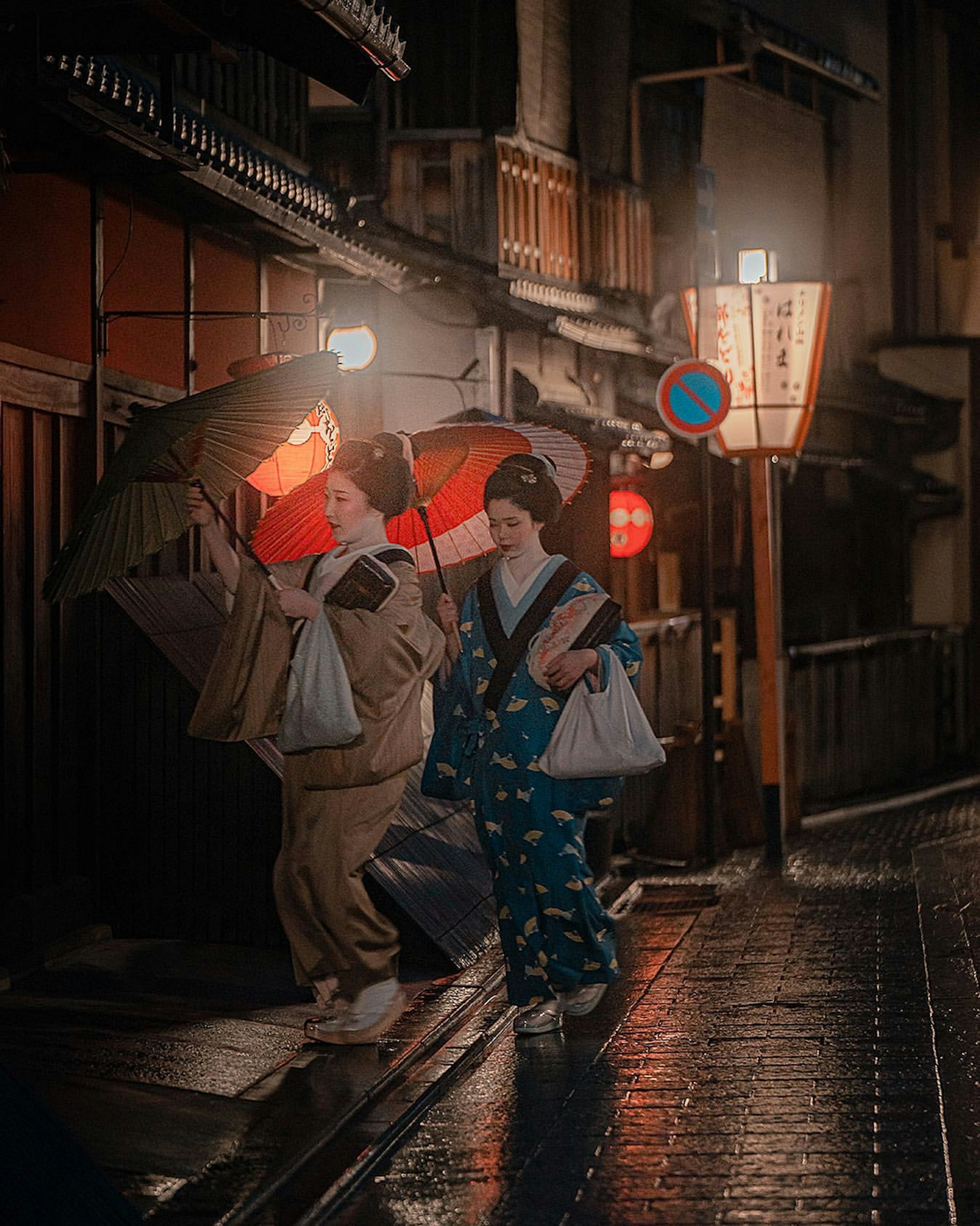 Zwei Frauen im Kimono, die nachts mit Regenschirmen gehen, beleuchtet von roten Laternen