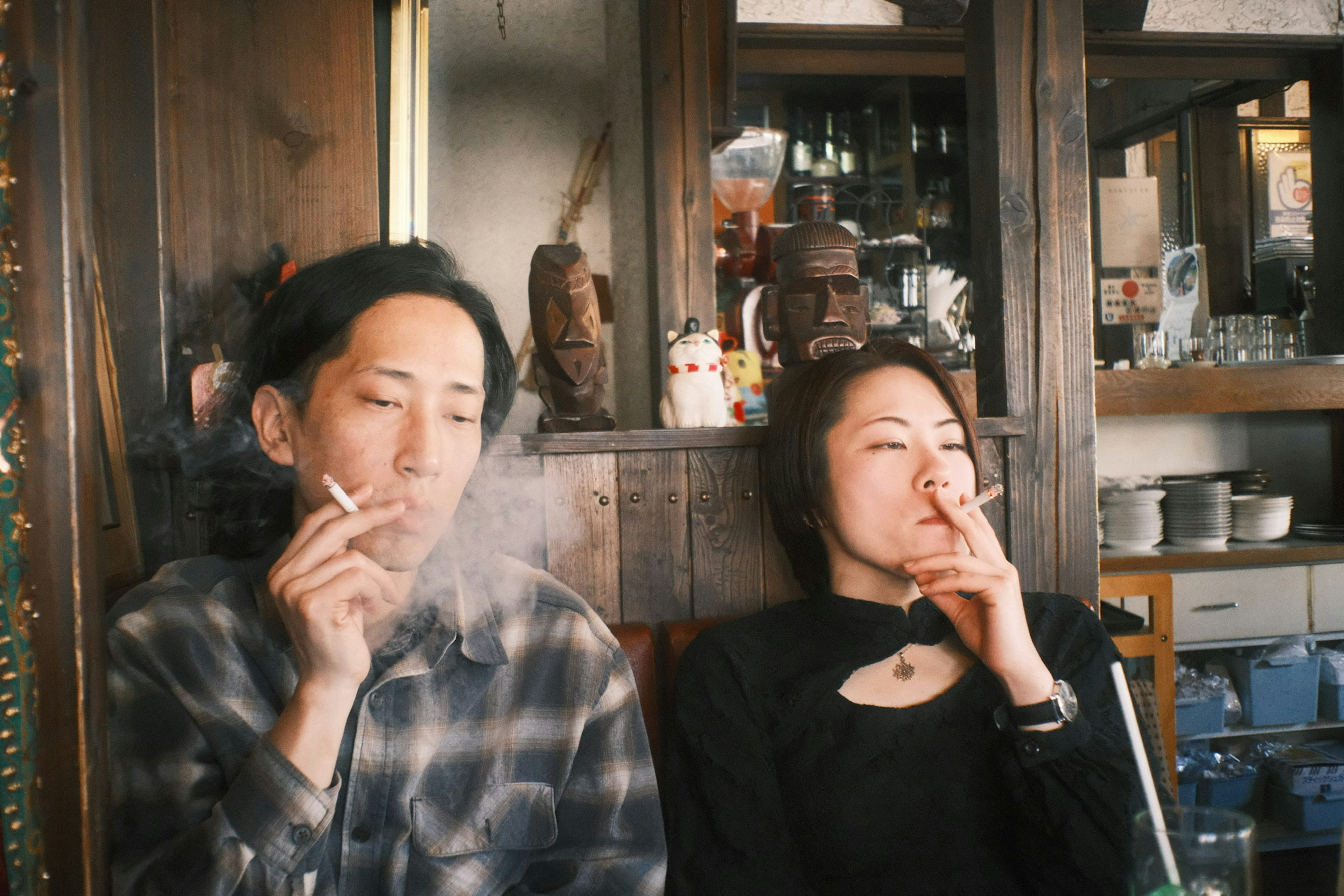 A man and woman smoking cigarettes in a cafe with wooden decor and drinks in the background