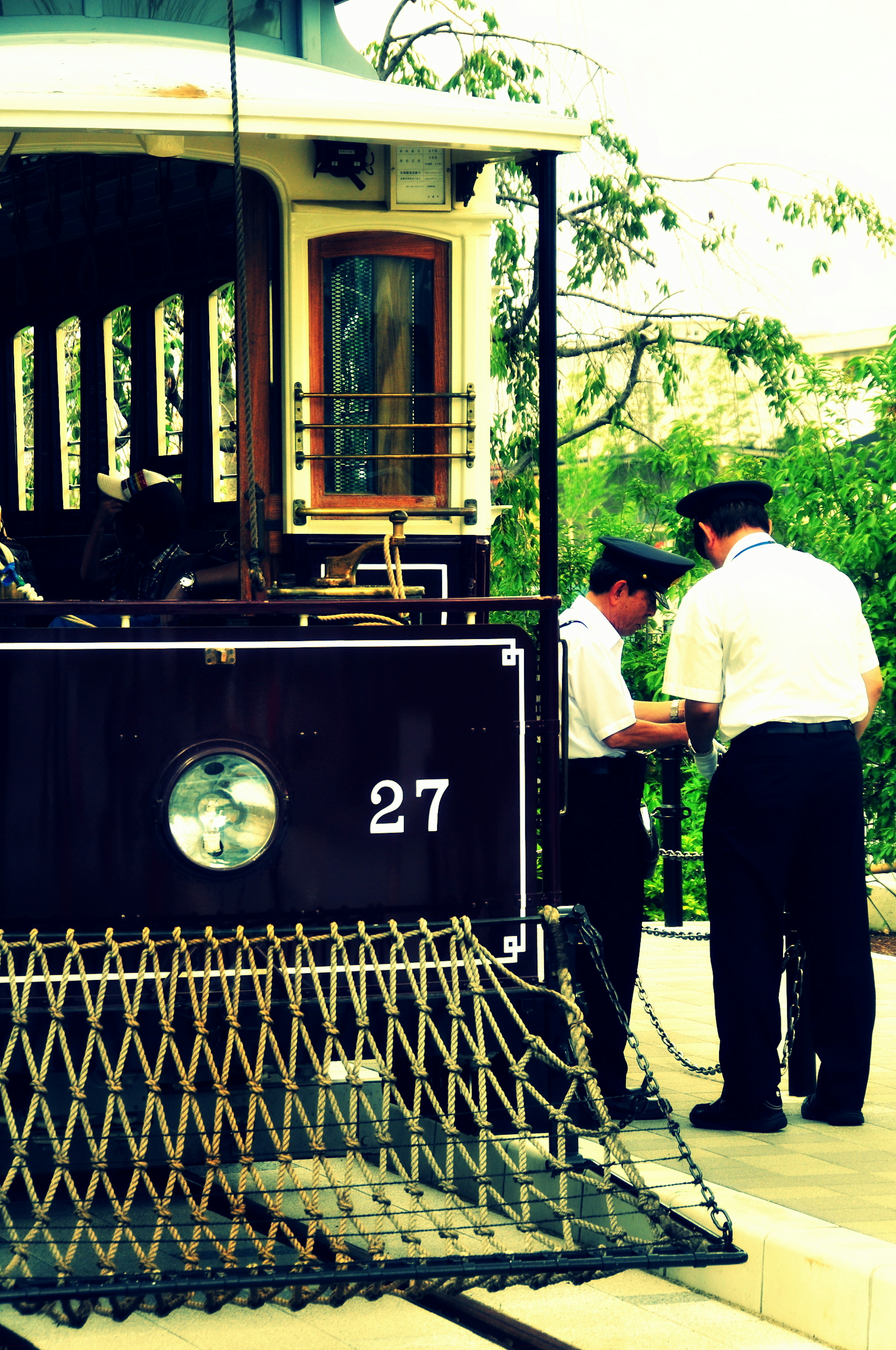 Two men working at the front of a trolley number 27 surrounded by green trees