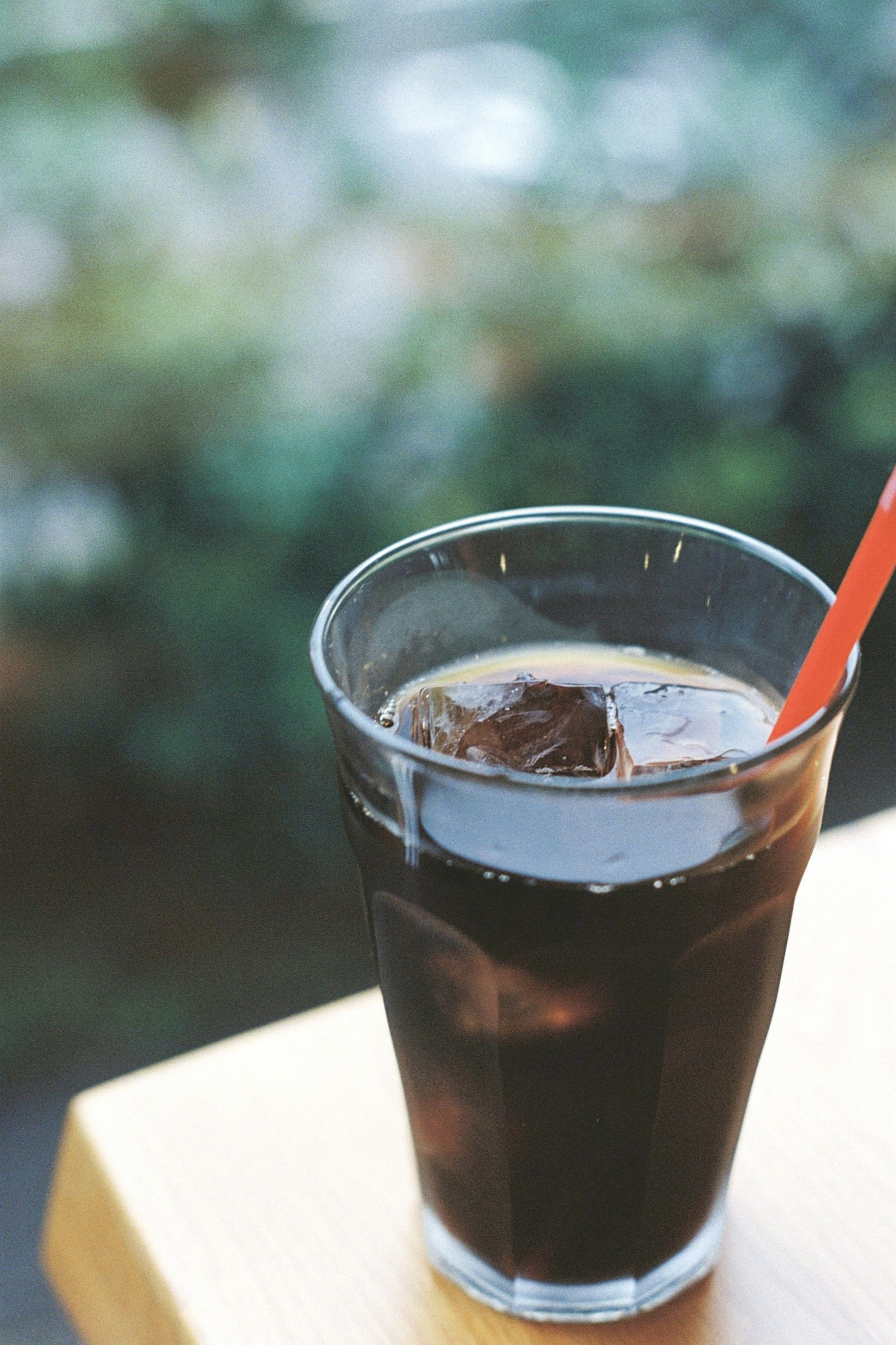 Un vaso transparente de café helado con cubitos de hielo y una pajita roja, rodeado de un fondo verde borroso