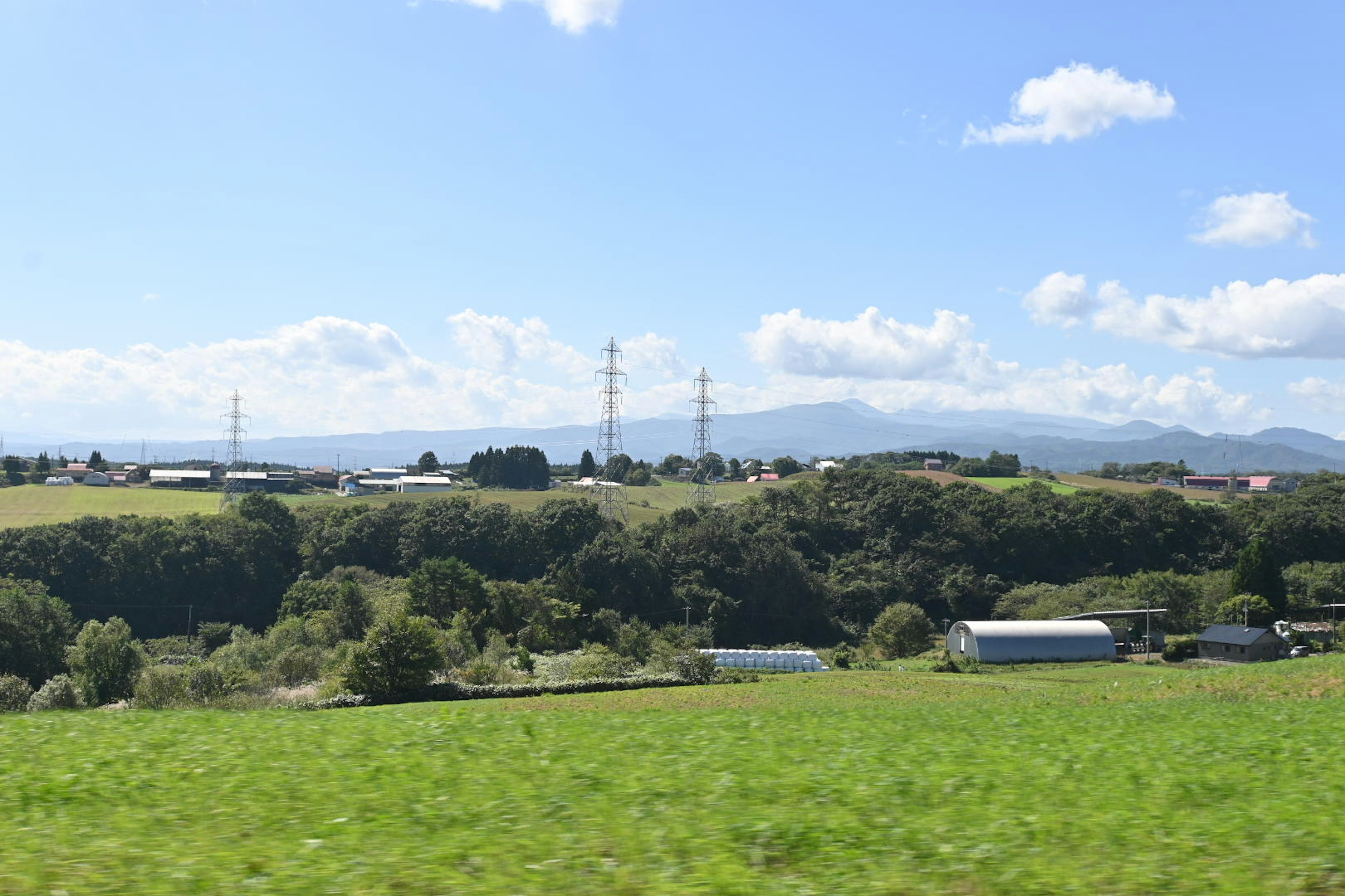 Rural landscape featuring green fields under a blue sky with power lines and distant mountains