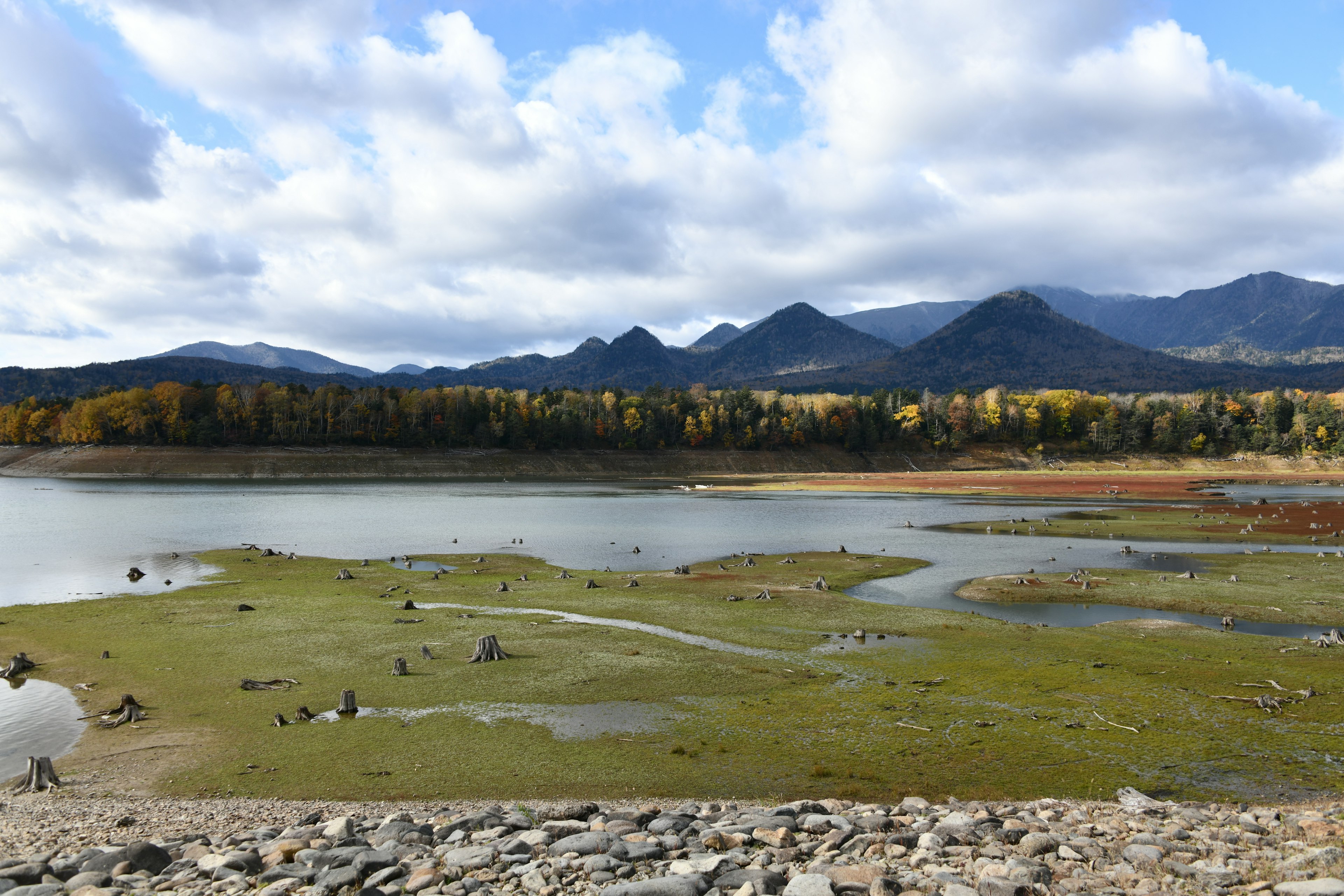 Scenic view of a lake and mountains with blue sky and clouds Grass and rocks scattered along the shore