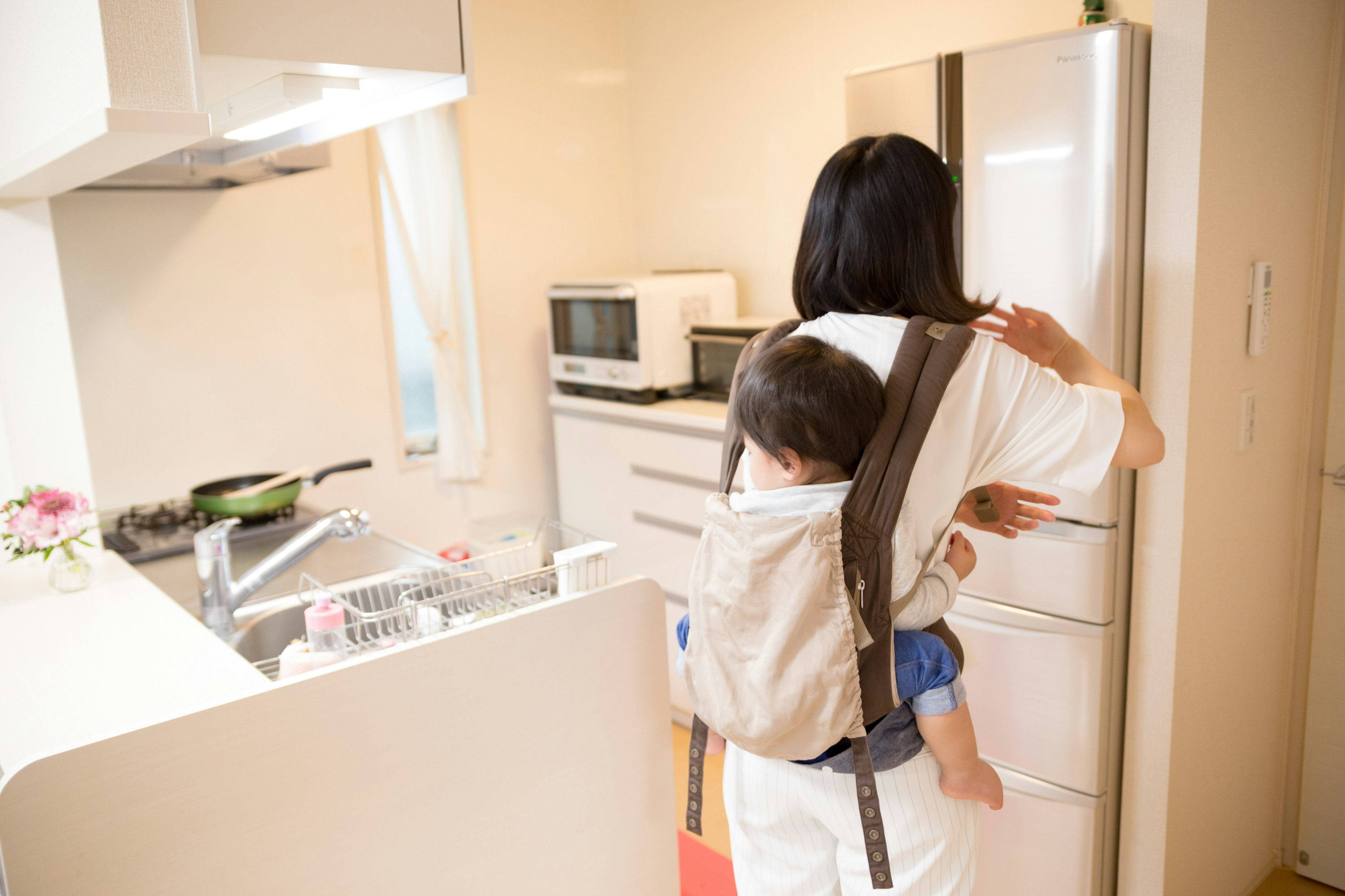 母親が子供を背負いながらキッチンで冷蔵庫を開けている