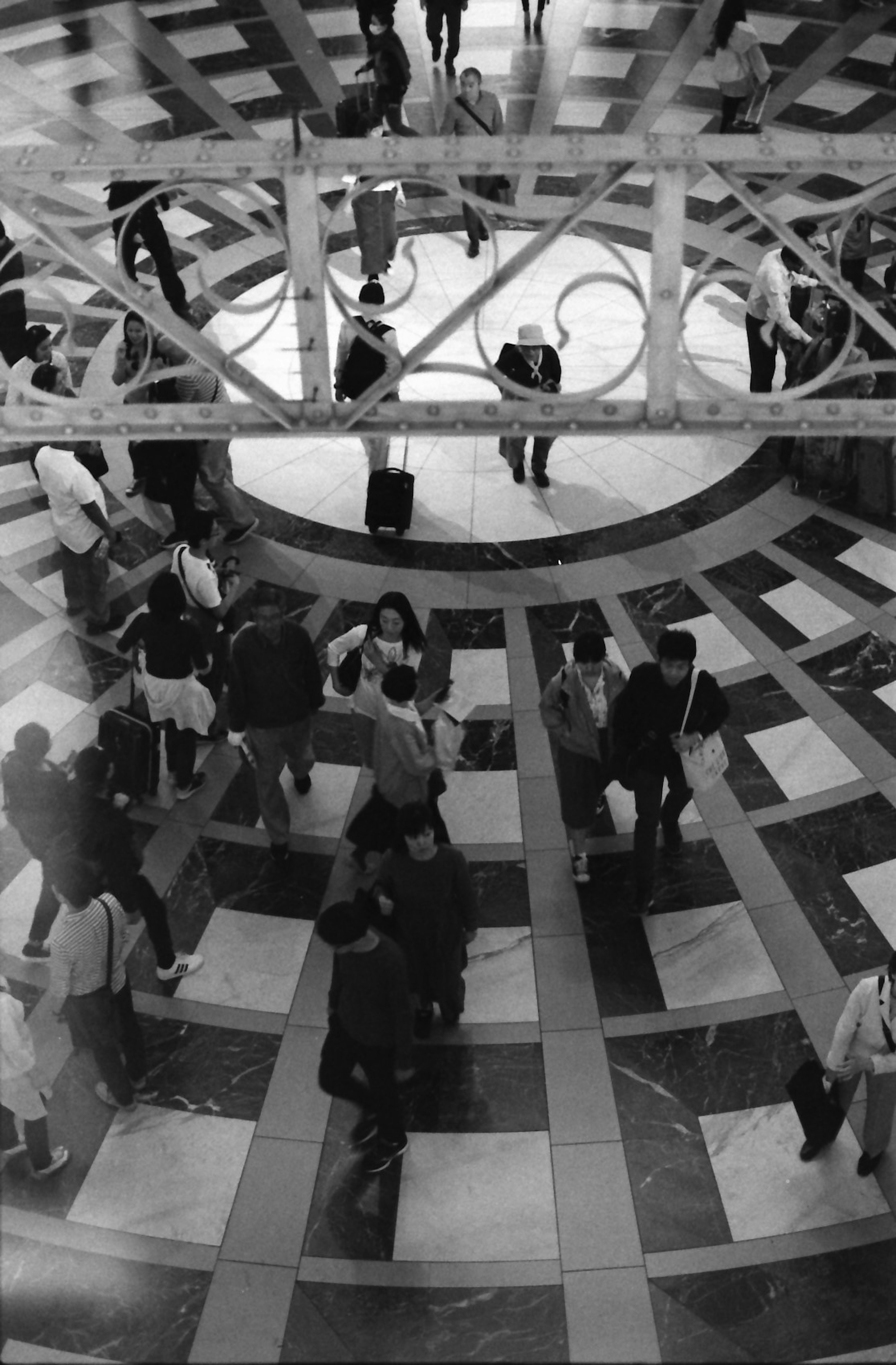 Multitud de personas caminando en una plaza de baldosas en blanco y negro