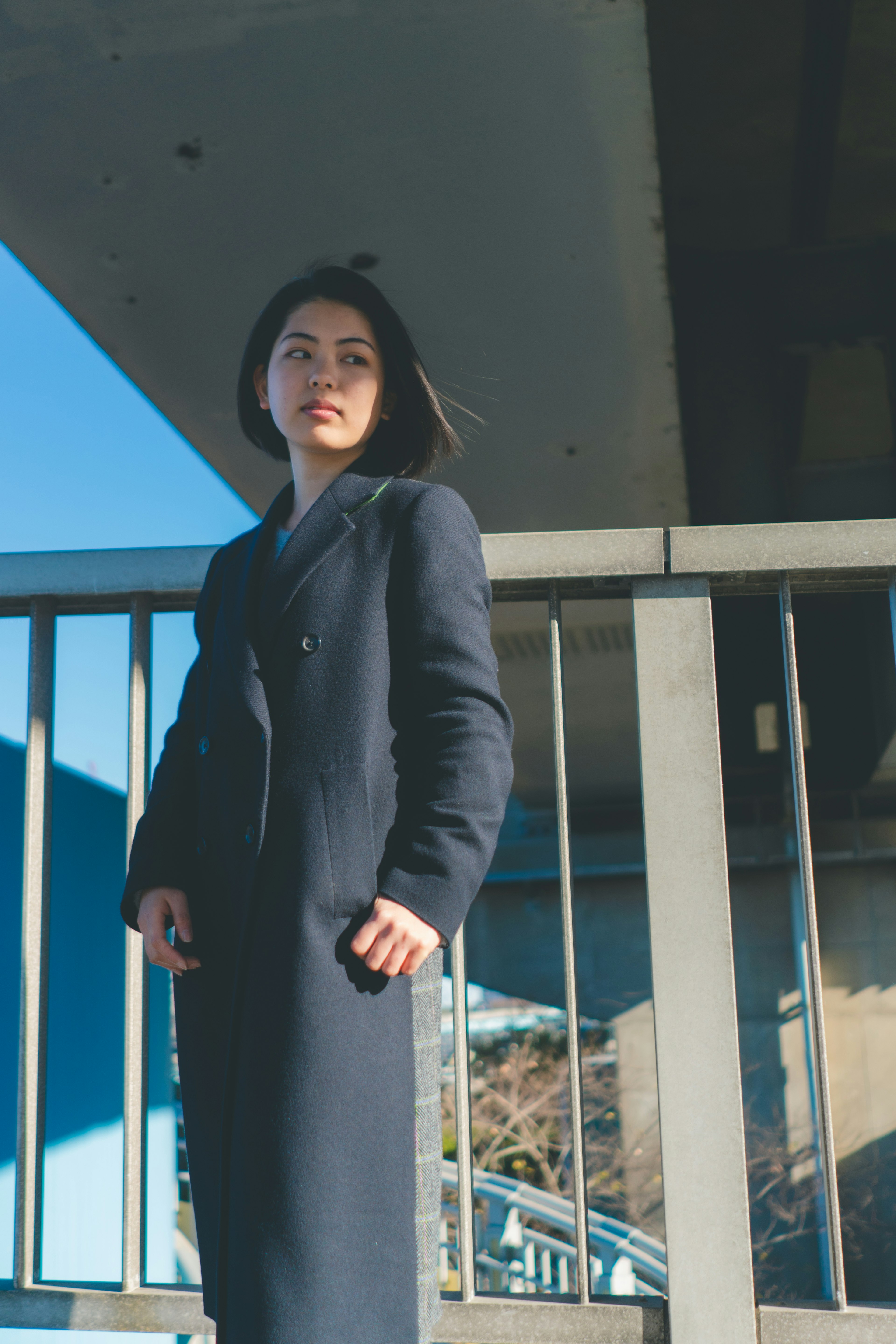 A woman in a black coat standing under a bridge with a clear blue sky