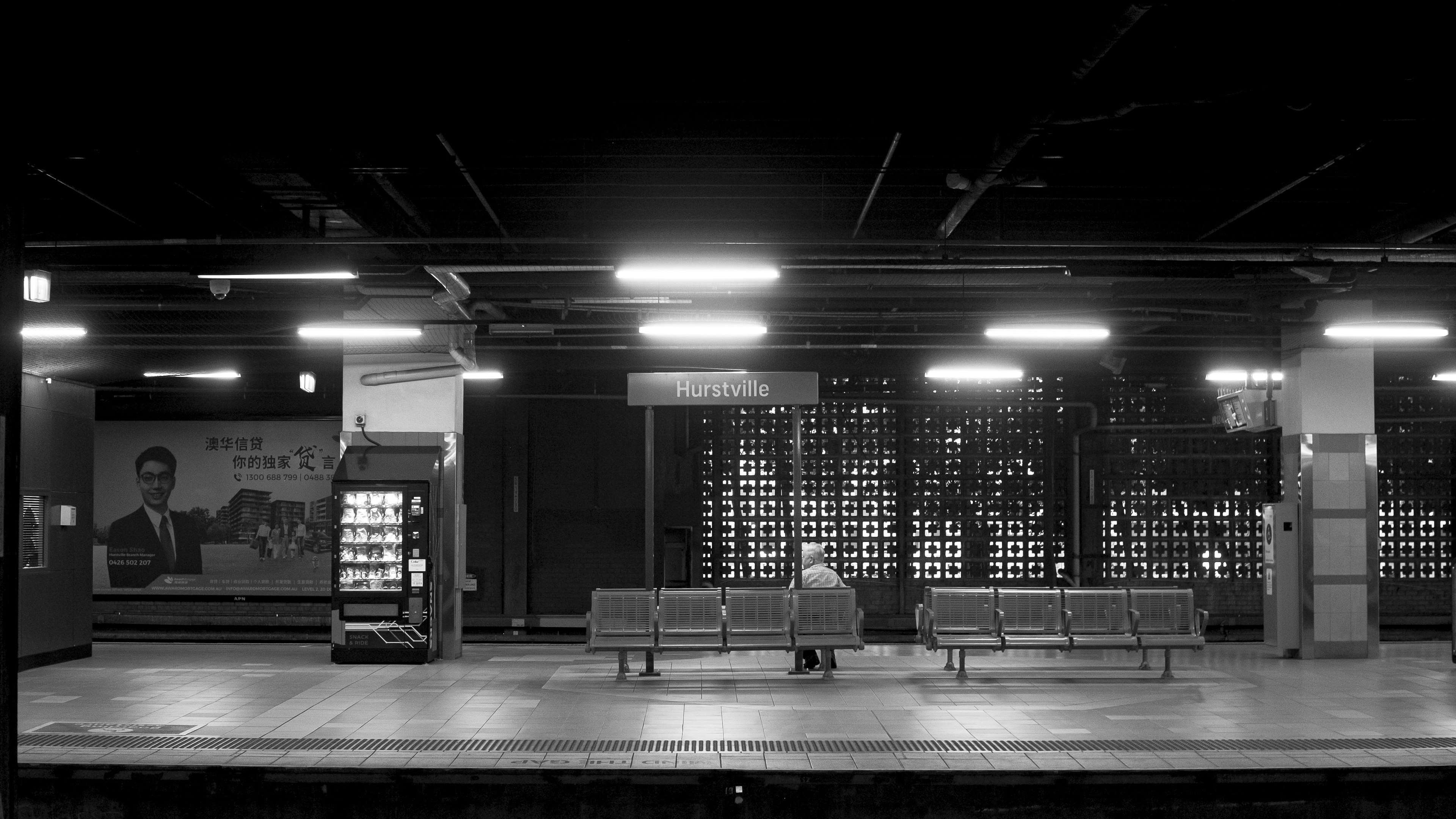 Black and white image of a quiet train platform featuring benches and a vending machine