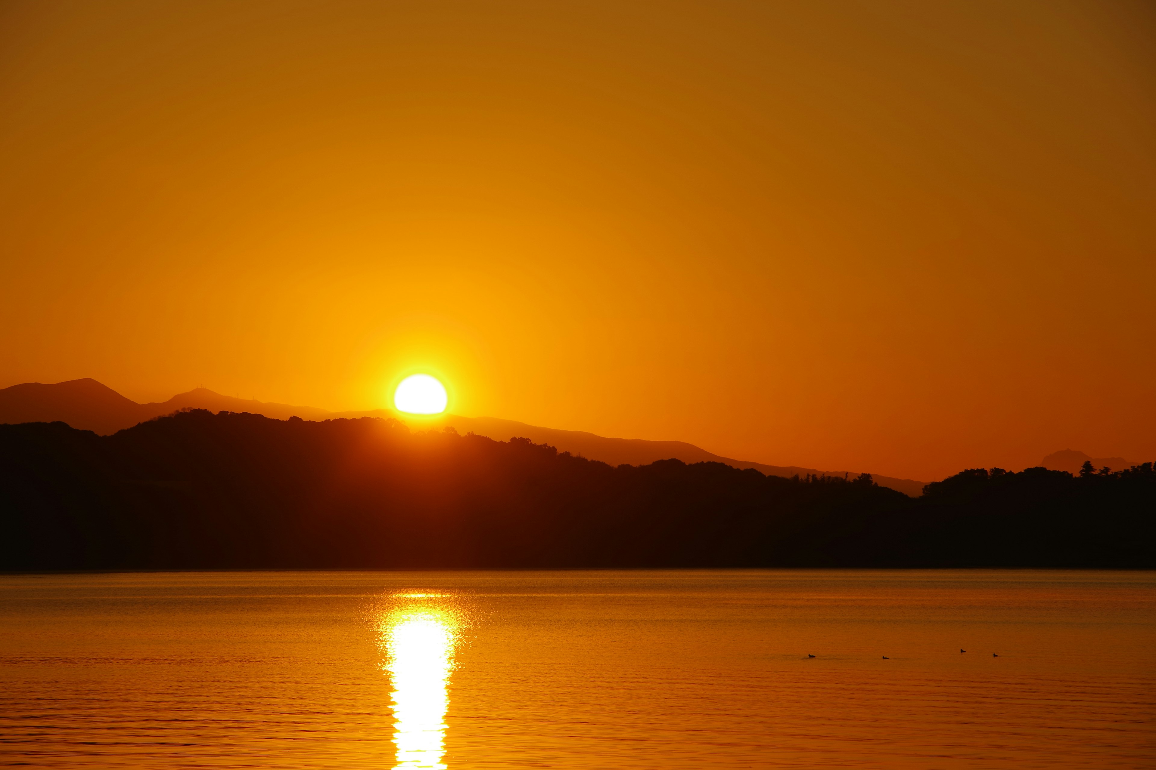 夕日が山の上に沈む様子と湖面に映るオレンジ色の反射