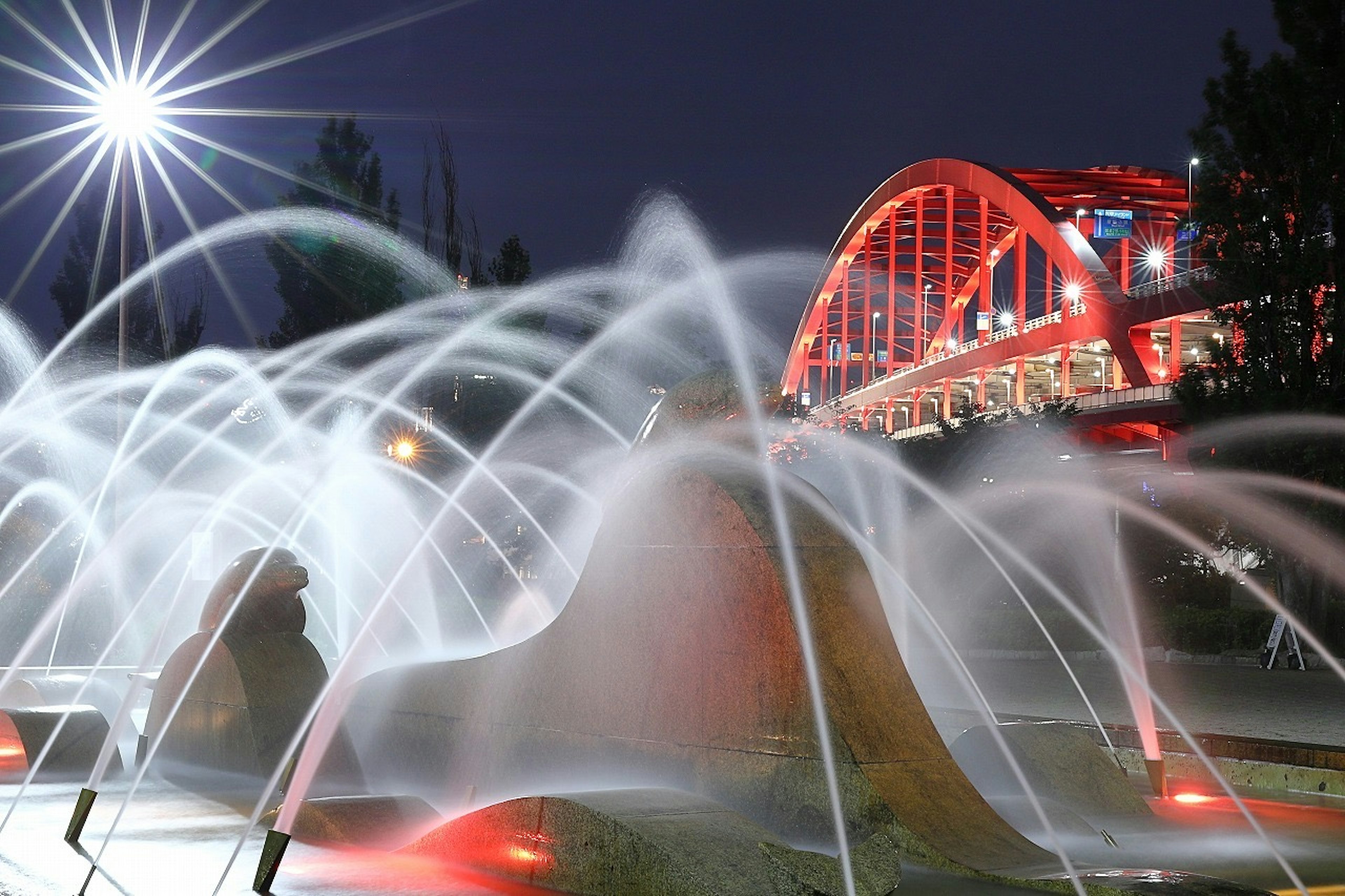 Fontaine illuminée dans un parc la nuit avec un pont en arc rouge